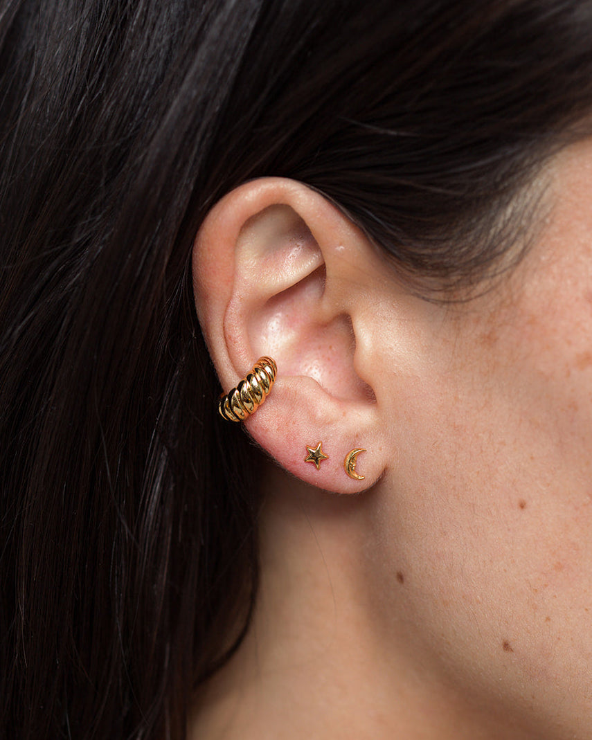 A close-up of a person’s ear adorned with three gold earrings: a thick twisted hoop in the upper cartilage, and two small celestial studs—a star and Mod + Jo’s Archie Moon Stud—in the lobe. The person has straight dark hair.