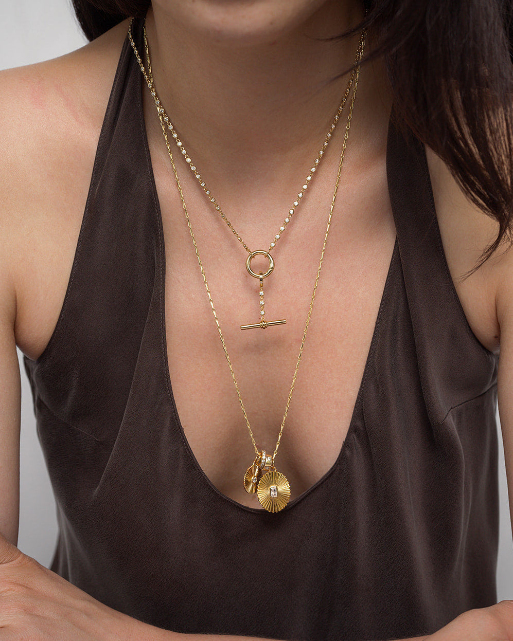 A woman wearing a deep brown top and two layered gold necklaces: a dainty chain necklace with a horizontal bar pendant, and the Mod + Jo Callum Tennis Charm Clip Necklace featuring a round, textured pendant with a small gold charm.