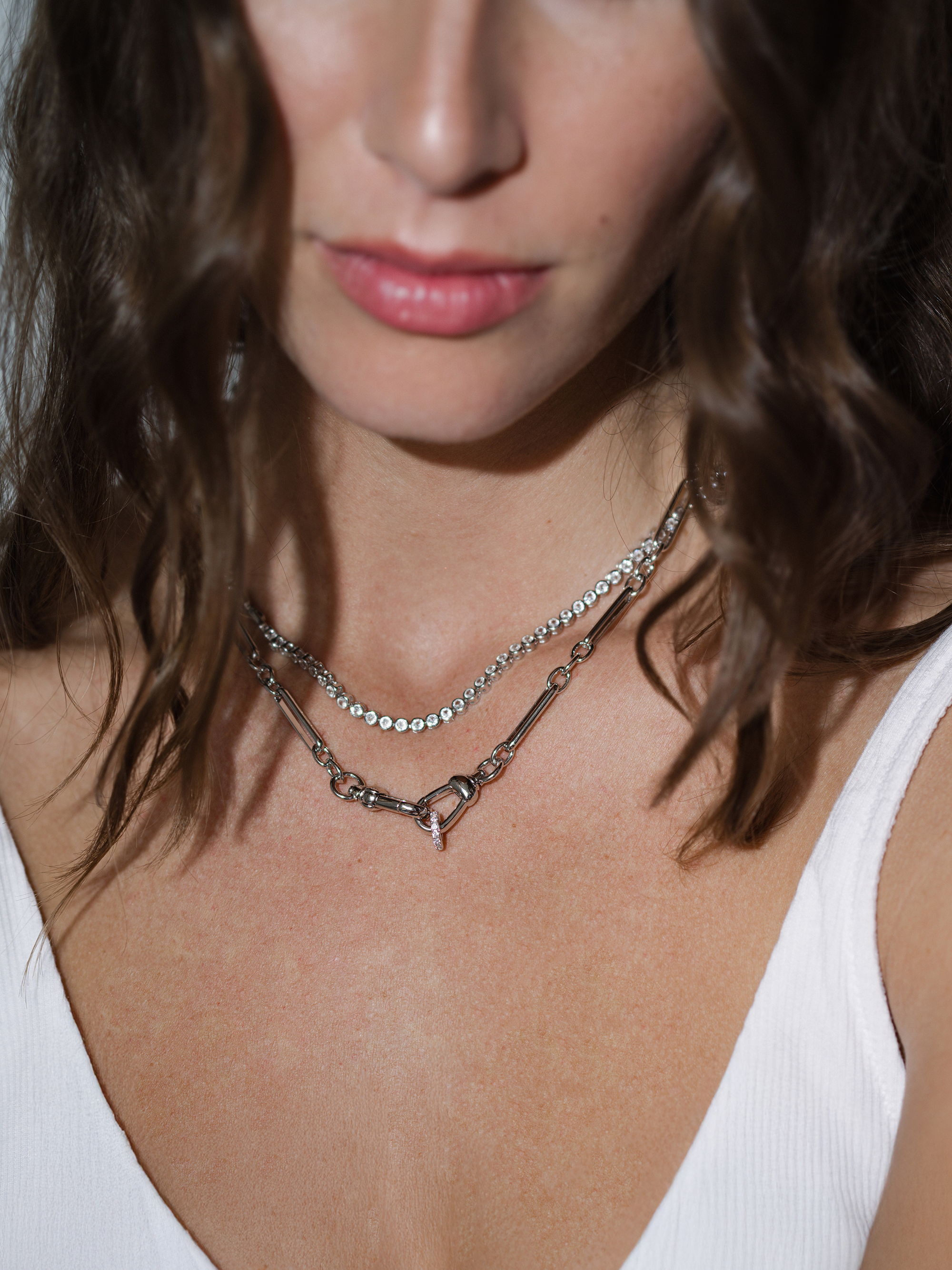 A woman with wavy brown hair wears a white top and two layered silver necklaces, one with small round stones and the other a simple chain with a clasp, partially visible against her bare skin.