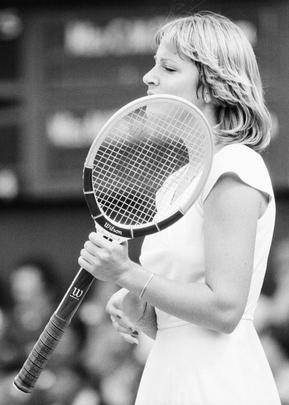 A woman in a white tennis outfit holds a tennis racket upright in front of her face during a match. The black-and-white image captures her mid-action, with a blurred background of spectators.