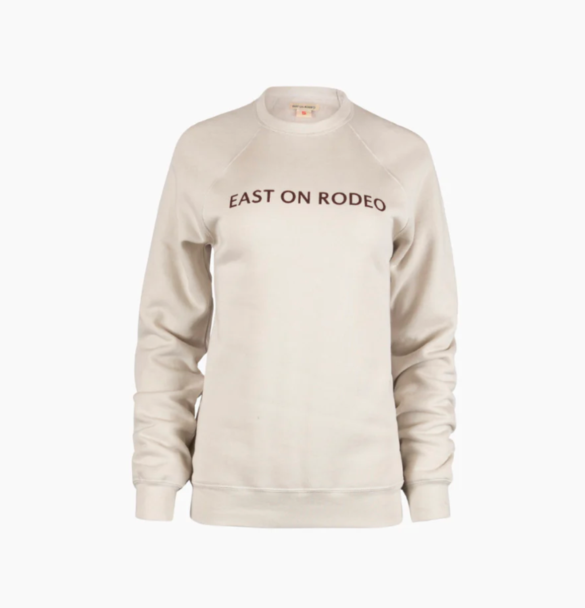 A light beige crewneck sweatshirt with the words EAST ON RODEO printed in capital letters across the chest. The sweatshirt has long sleeves and ribbed cuffs and hem.