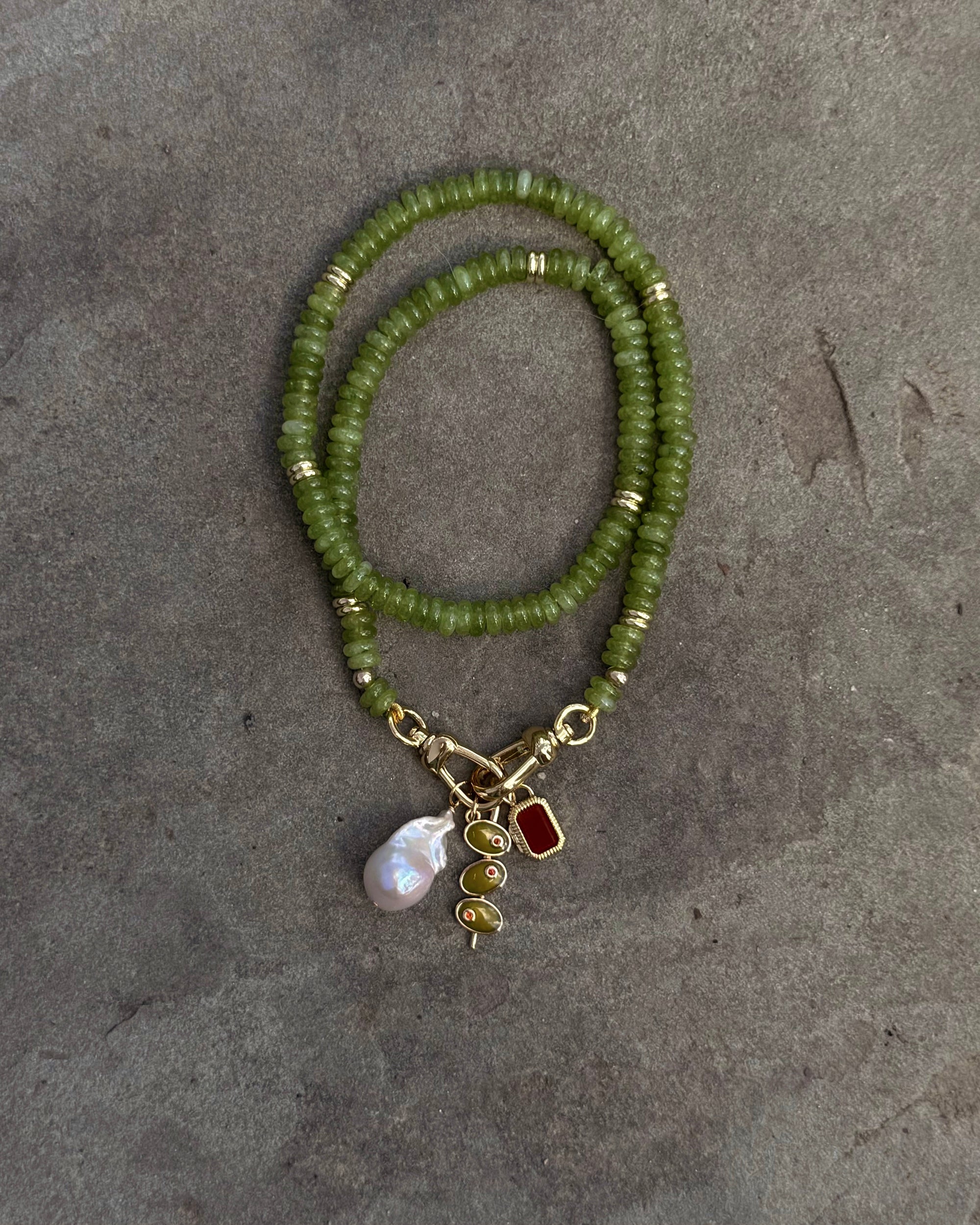 The Sonia Gemstone Necklace by Mod + Jo, crafted with genuine gemstone beads and gold accents, is displayed on a gray background. It features three stunning charms: a pearl, a gold spiral, and a red gem—ideal for handmade jewelry lovers.