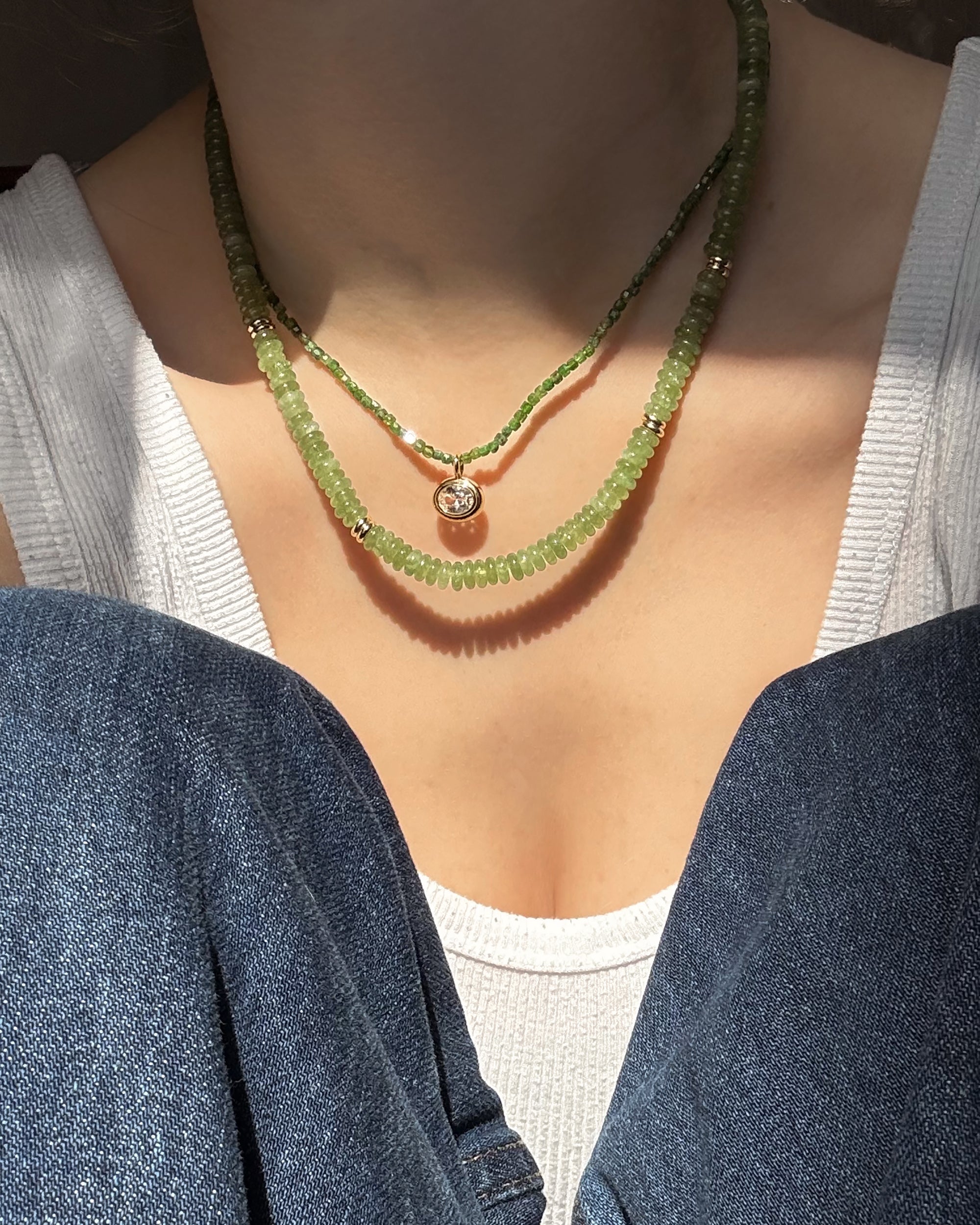A person in a white ribbed tank top and blue jeans sits with knees up, layering two Mod + Jo Merit Gemstone Necklaces—hand-crafted green pieces, one accented by a small round pendant—around their neck.