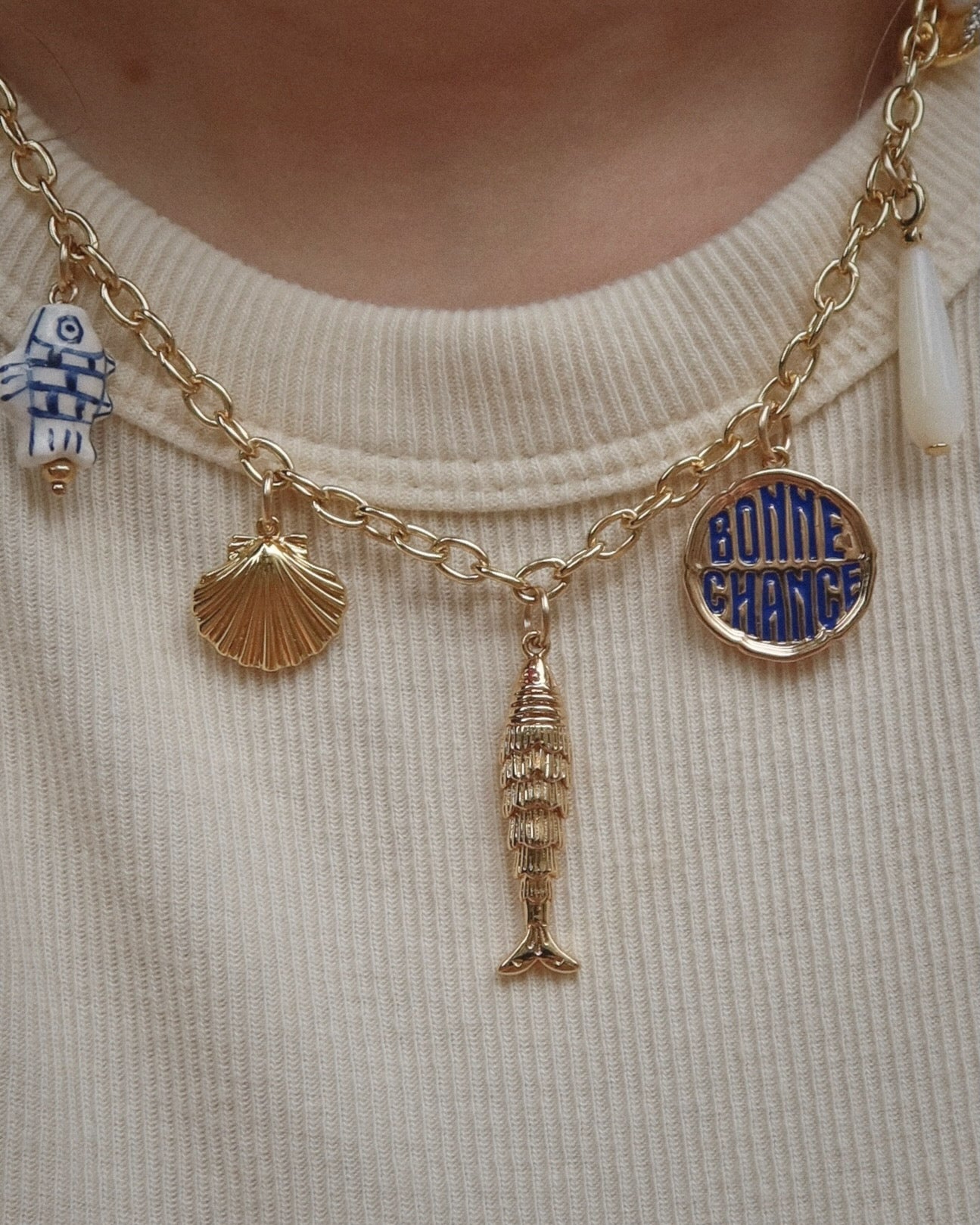 A gold charm necklace with five pendants: a blue-and-white fish, a gold seashell, a gold fish, a round blue-and-gold BONNE CHANCE charm, and a white teardrop, worn over a beige ribbed shirt.