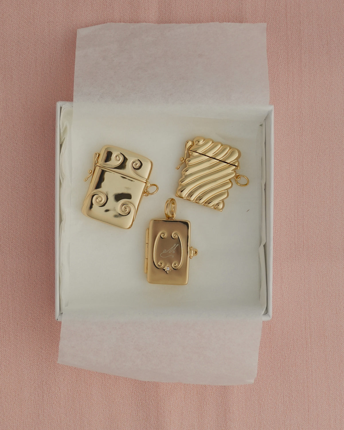 A set of Caspia Vesta Case Charms in a white gift box, featuring three gold-plated metal charms: one rectangular piece with a small crystal, one with a wavy texture, and one with a swirl design.
