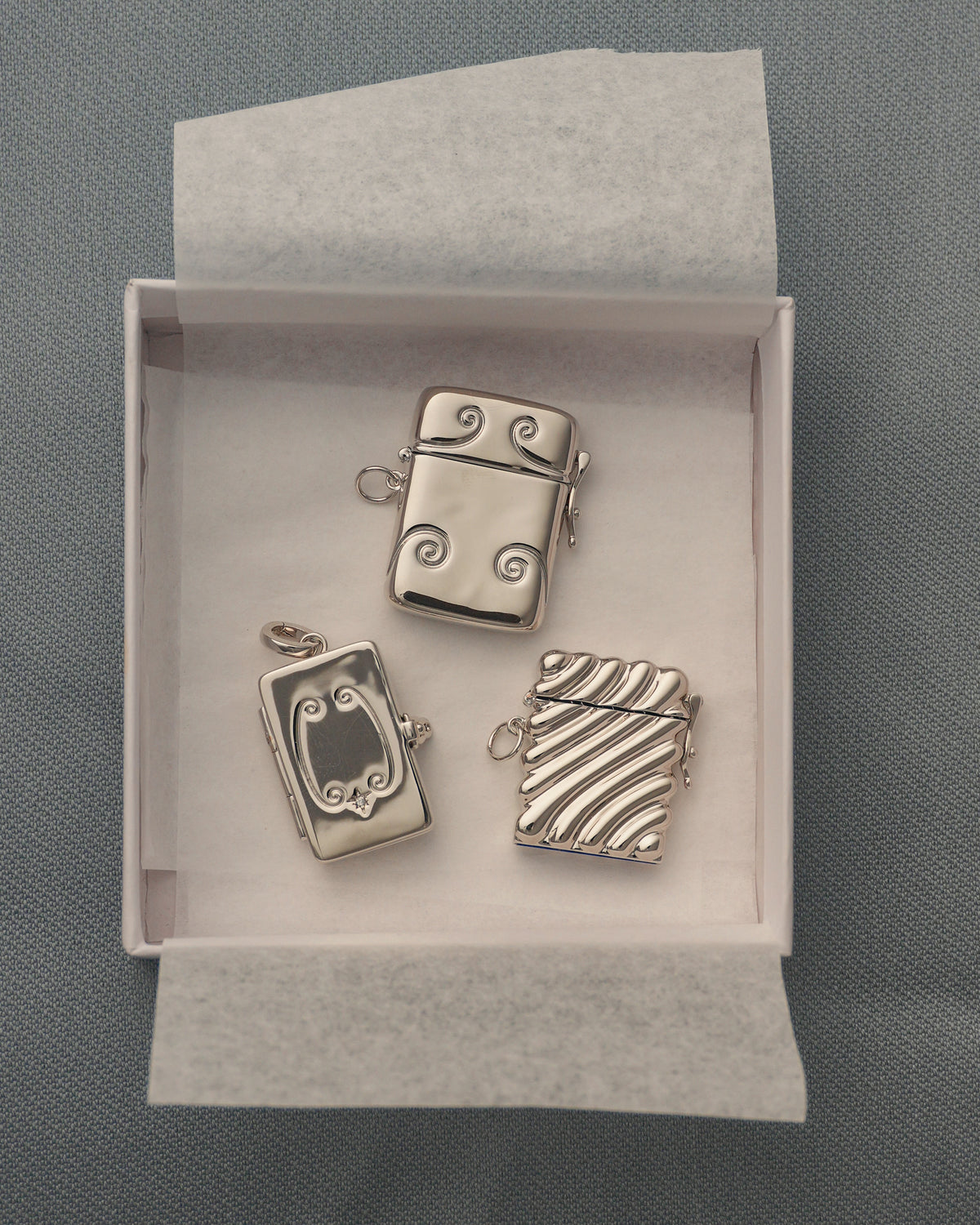Three small, silver-plated Delphine Locket charms in assorted shapes and decorative patterns, including a rectangular locket, nestled inside an open white gift box.