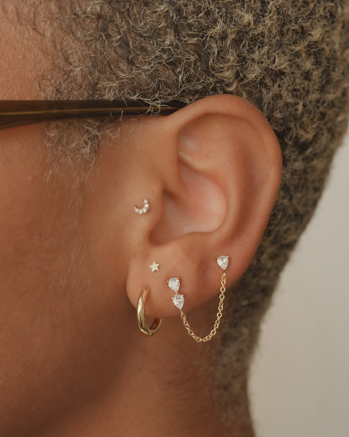 Close-up of an ear wearing the Reneé + Tatum Set, featuring gold earrings including a small moon stud, a star stud, polished hoops, and a double-pear-shaped stone earring connected by a delicate chain.