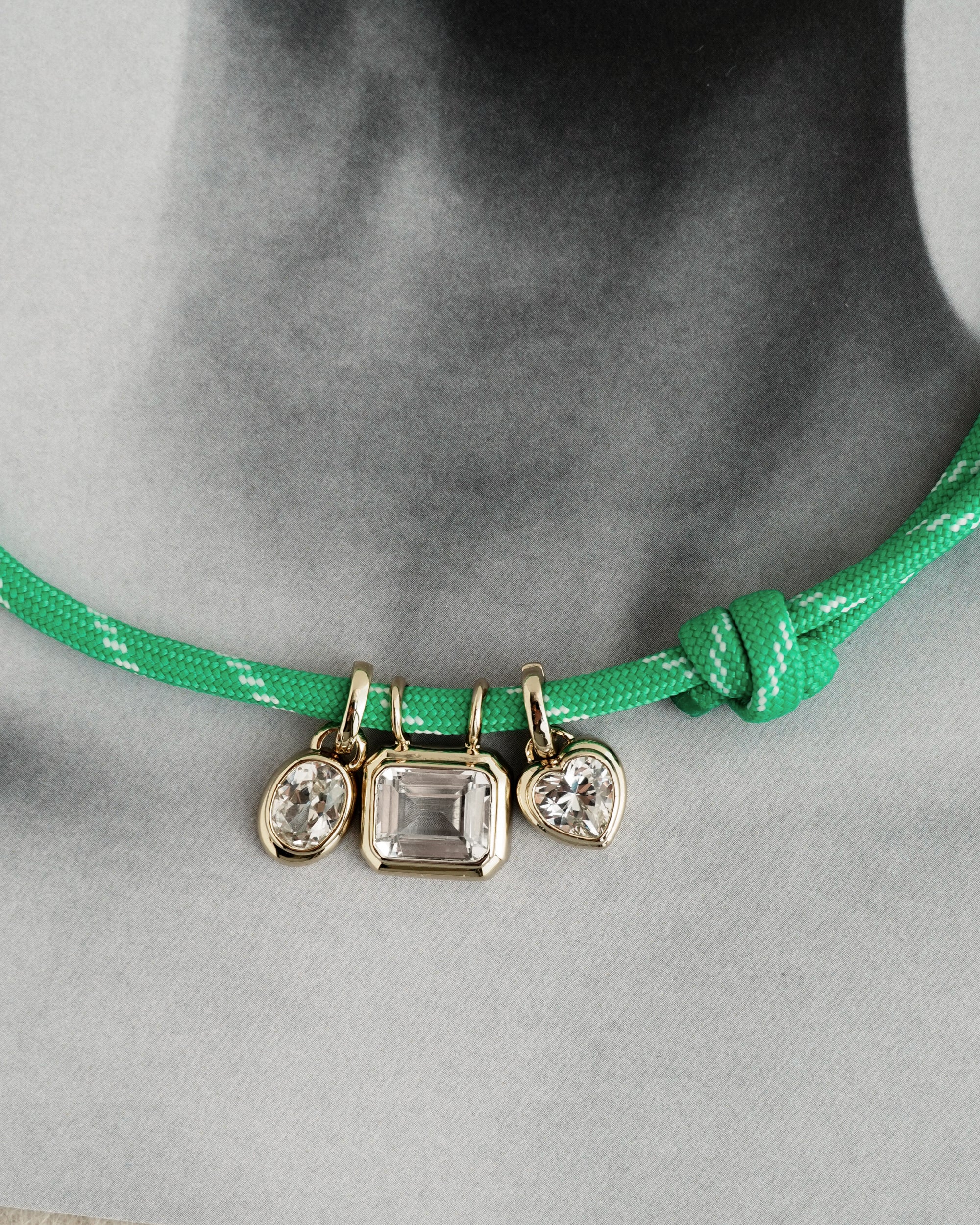 The *Preorder* Tommy Sapphire Pendant Necklace by Mod + Jo is a green adjustable paracord necklace with a knot and three gemstone charms—including a lab-grown white sapphire—set in gold, shown on a grayscale photo of a neck.