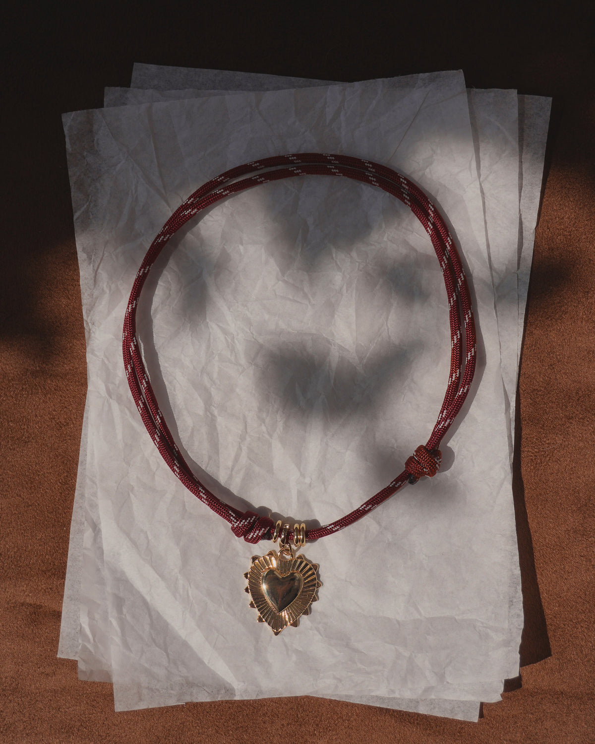 The Tommy Cord Necklace by Mod + Jo, featuring a red paracord design with a gold heart pendant and subtle gold plate accents, rests on crumpled white paper as sunlight and shadows create a warm, textured backdrop.