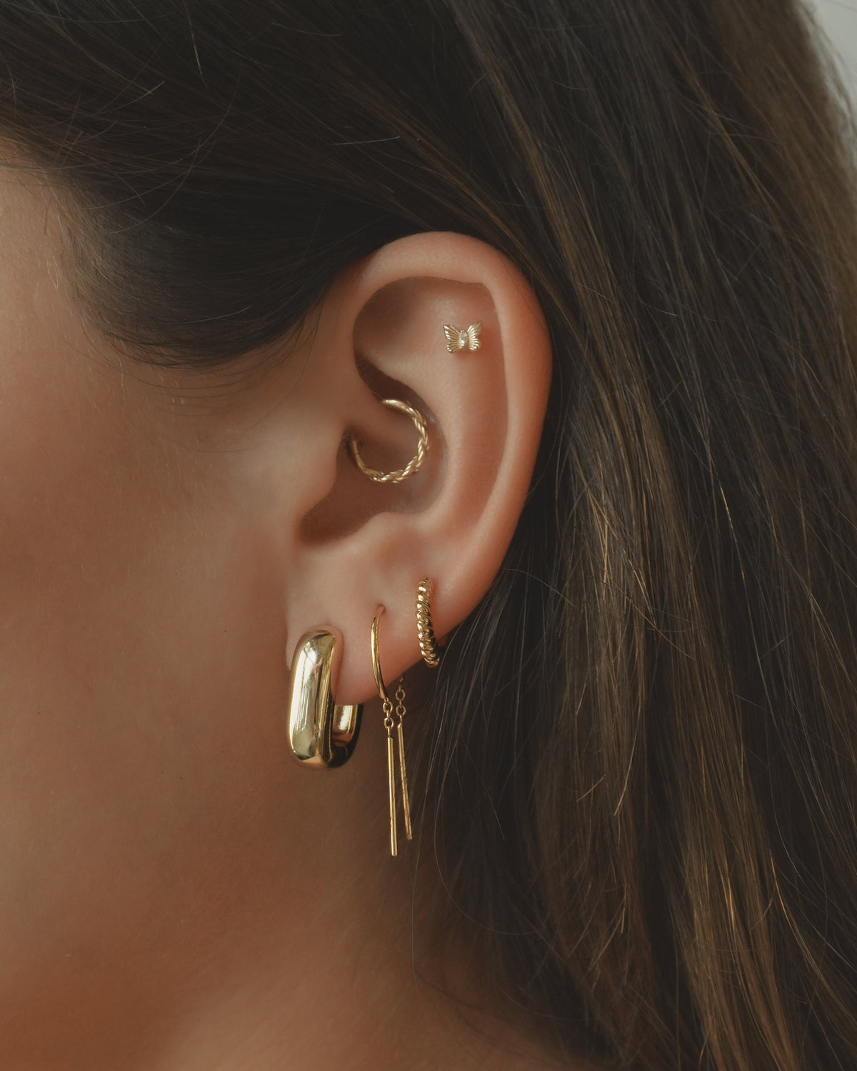 A close-up of a woman’s ear showcases multiple gold earrings from Mod + Jo, including the Mila Smooth Threader Huggies, alongside a large 14k gold hoop, a small hoop, a cubic zirconia-accented bar earring, a butterfly stud, and a twisted cuff. Her brown hair is partially visible.