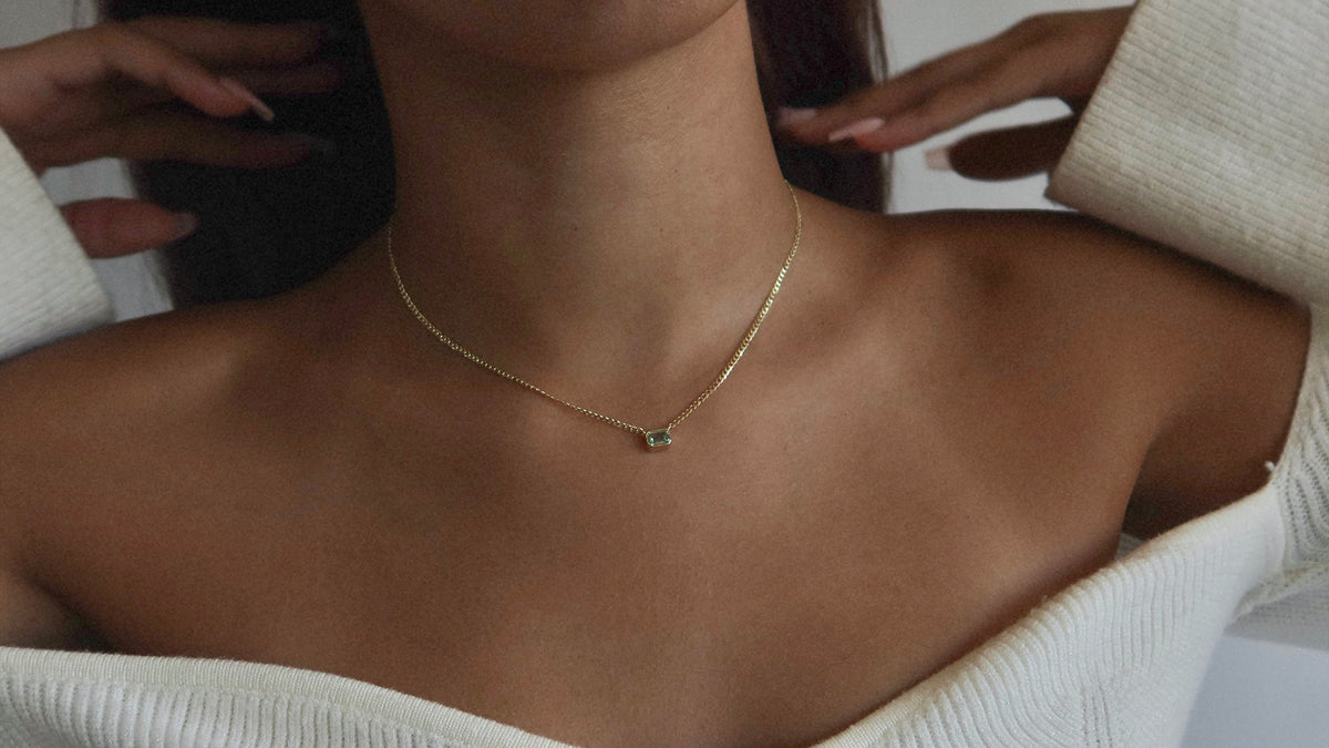 A woman wearing an off-the-shoulder white sweater displays a delicate gold necklace with a small green stone pendant resting at her collarbone. Her hands are raised near her neck.