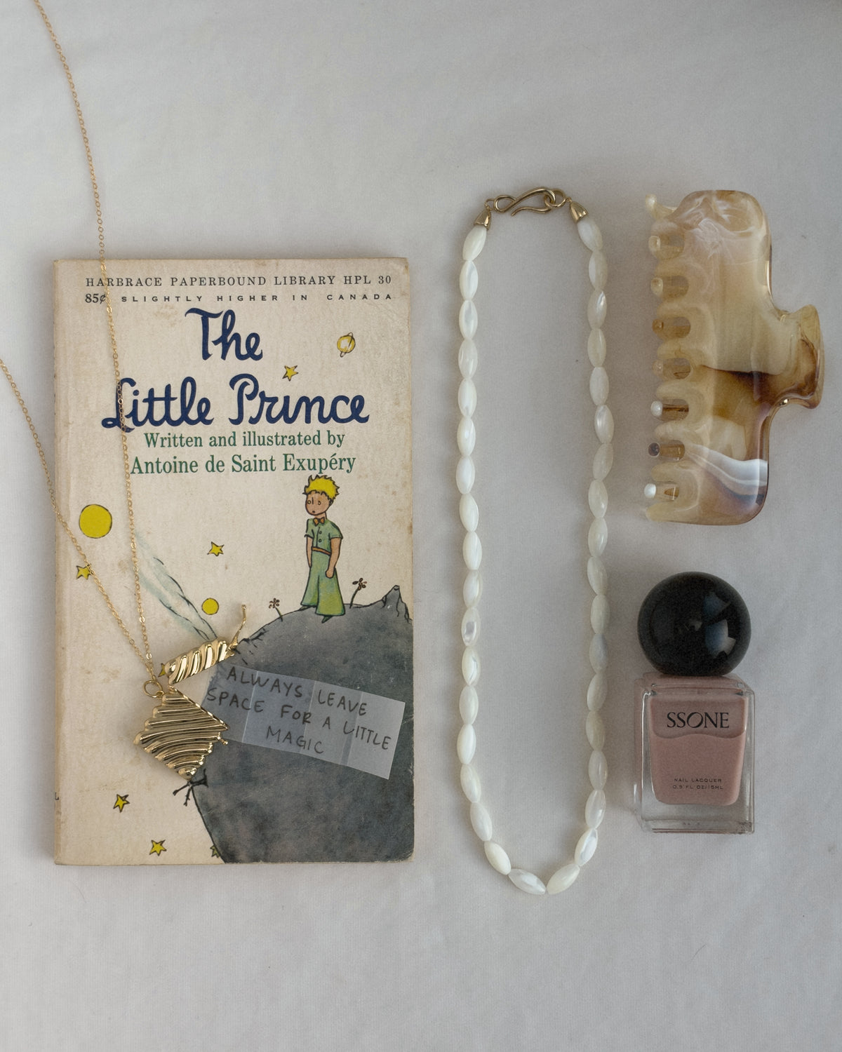 A Mod + Jo Sutton Gemstone Necklace, a mother of pearl bead necklace, a marble-patterned hair claw, and a bottle of neutral nail polish arranged next to a copy of The Little Prince.