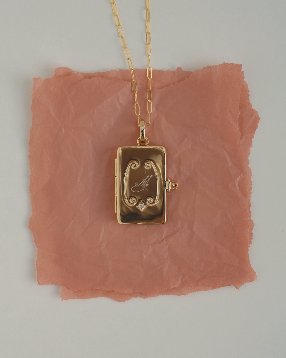 Delphine Locket Charm: a gold-plated rectangular locket on a delicate chain, featuring an engraved letter M and a small embedded cubic zirconia.