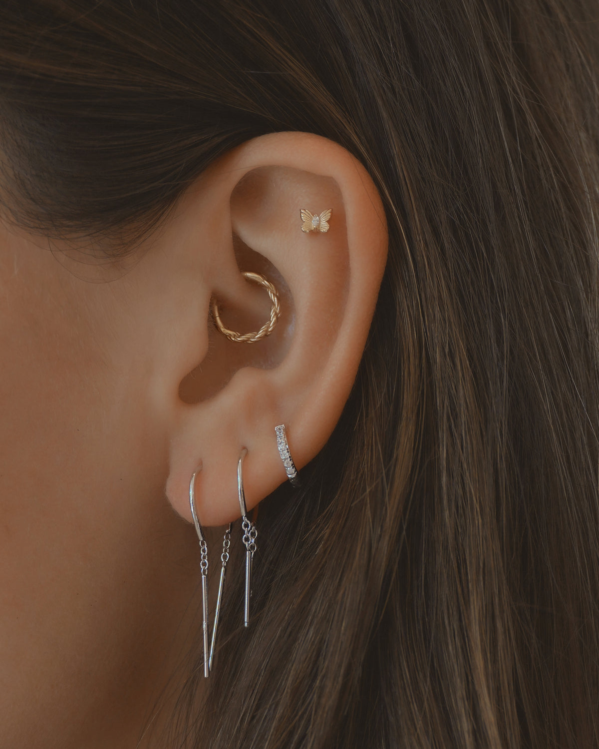 Close-up of an ear adorned with four earrings, including Mila Smooth Threader Huggies by Mod + Jo, a gold butterfly stud, a gold twisted hoop, and a rhodium-plated silver hoop embellished with cubic zirconia accents. Two of the earrings feature delicate dangling silver chains. Brown hair is partially visible in the background. These hypoallergenic pieces combine style and comfort.