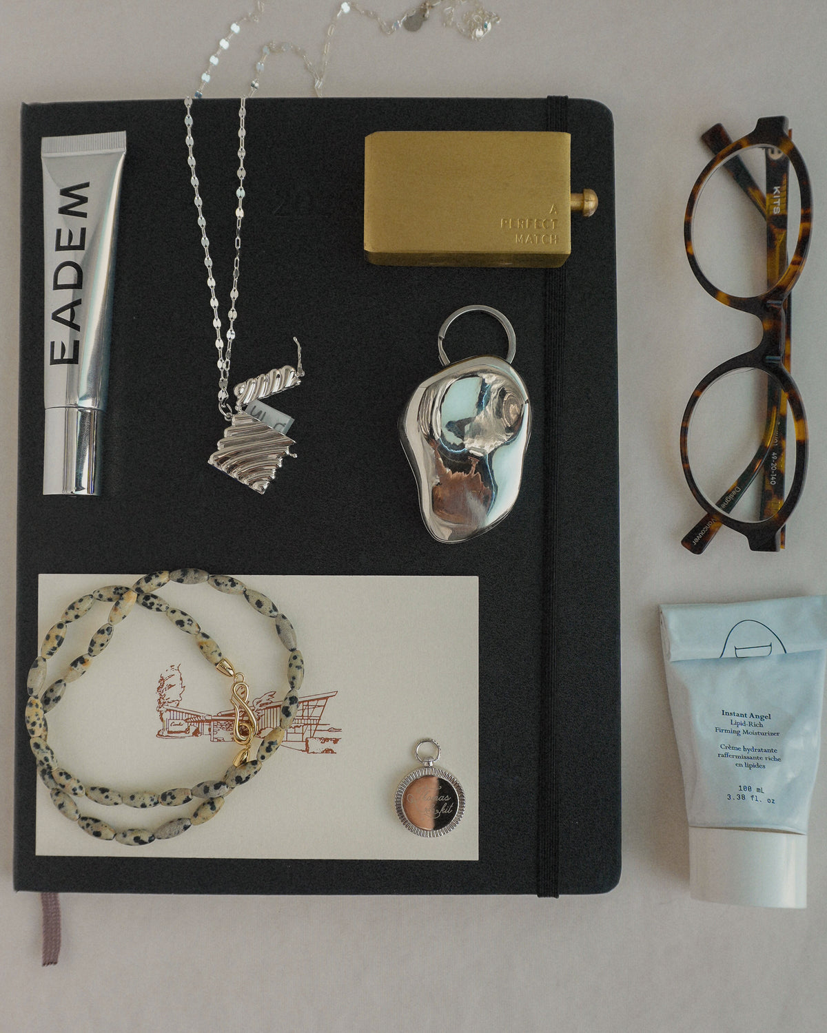 A flat lay of a black notebook surrounded by a Sutton Gemstone Necklace, a metal chain necklace, a gold-toned lighter, tortoiseshell glasses, and two tubes of skincare.
