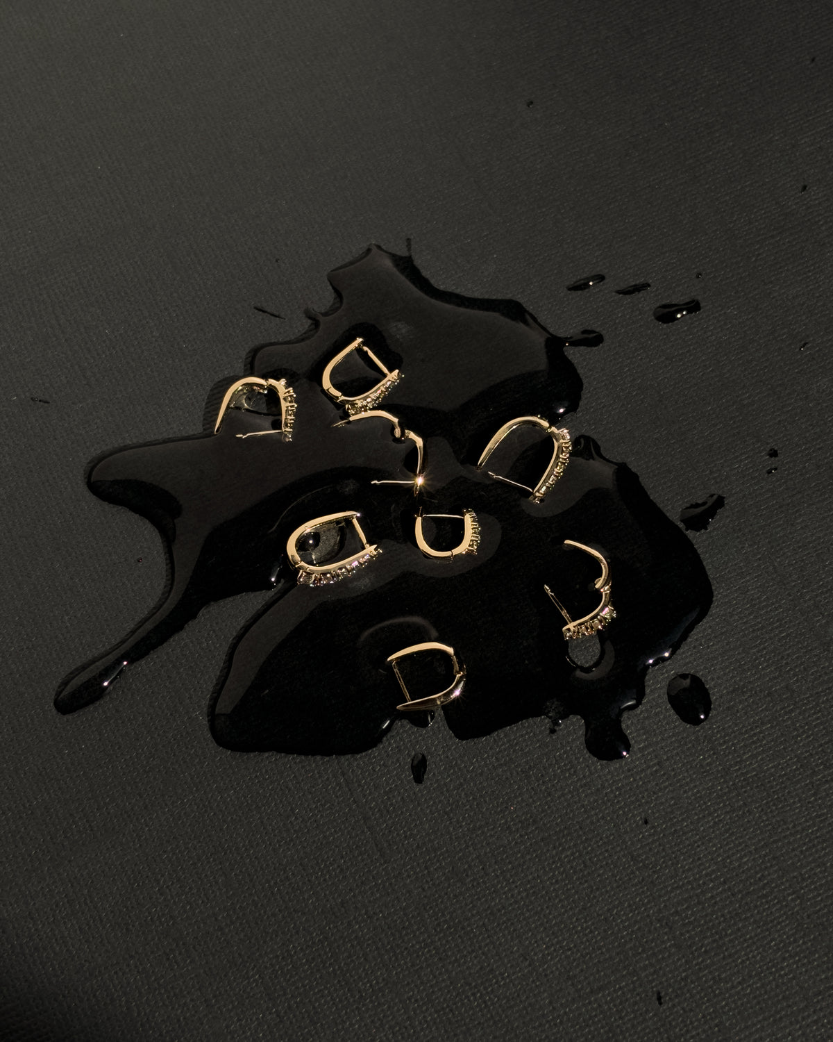 Six 14k gold plated Jules Medium Huggies, some featuring small cubic zirconia inset stones, resting on a black liquid puddle.
