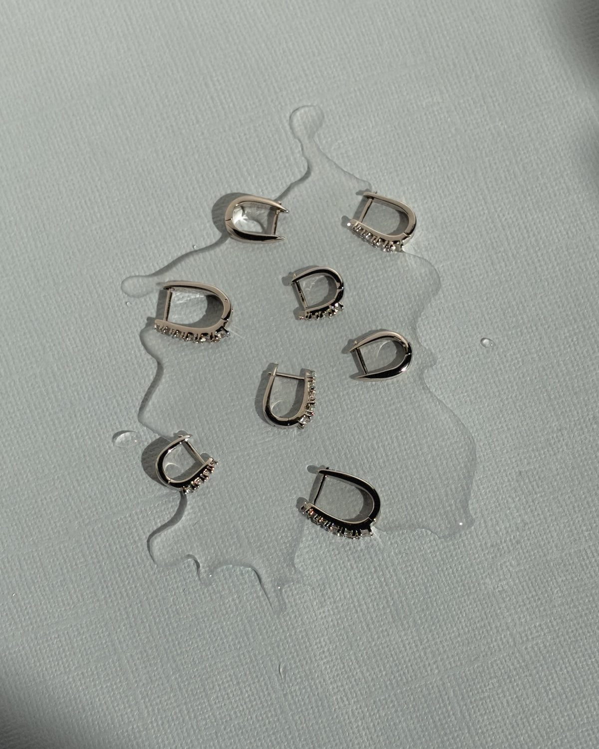Six Jules Grande Huggies tarnish-resistant silver hoop earrings with cubic zirconia accents arranged on a light grey surface with a small pool of water.