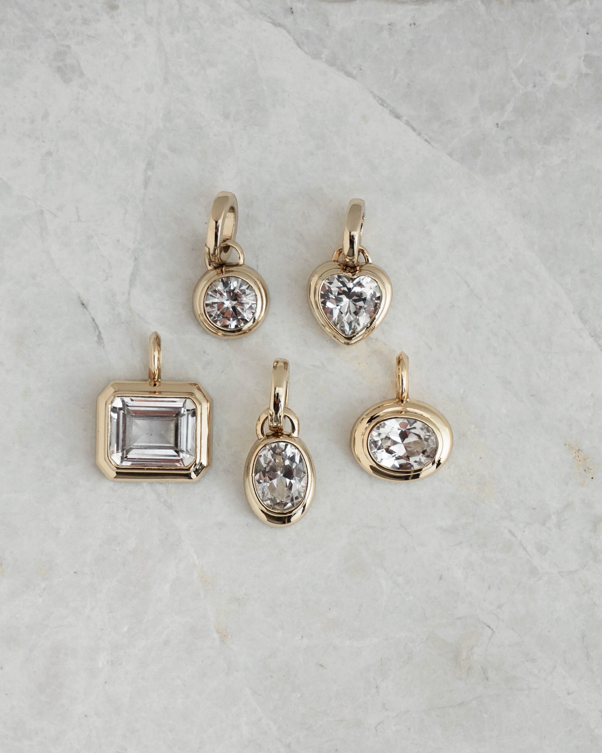 Five gold pendants—including Mod + Jo's Elsie Sapphire Bezel Charm—highlight shapes like circle, heart, oval, and rectangle on a light gray marble surface.