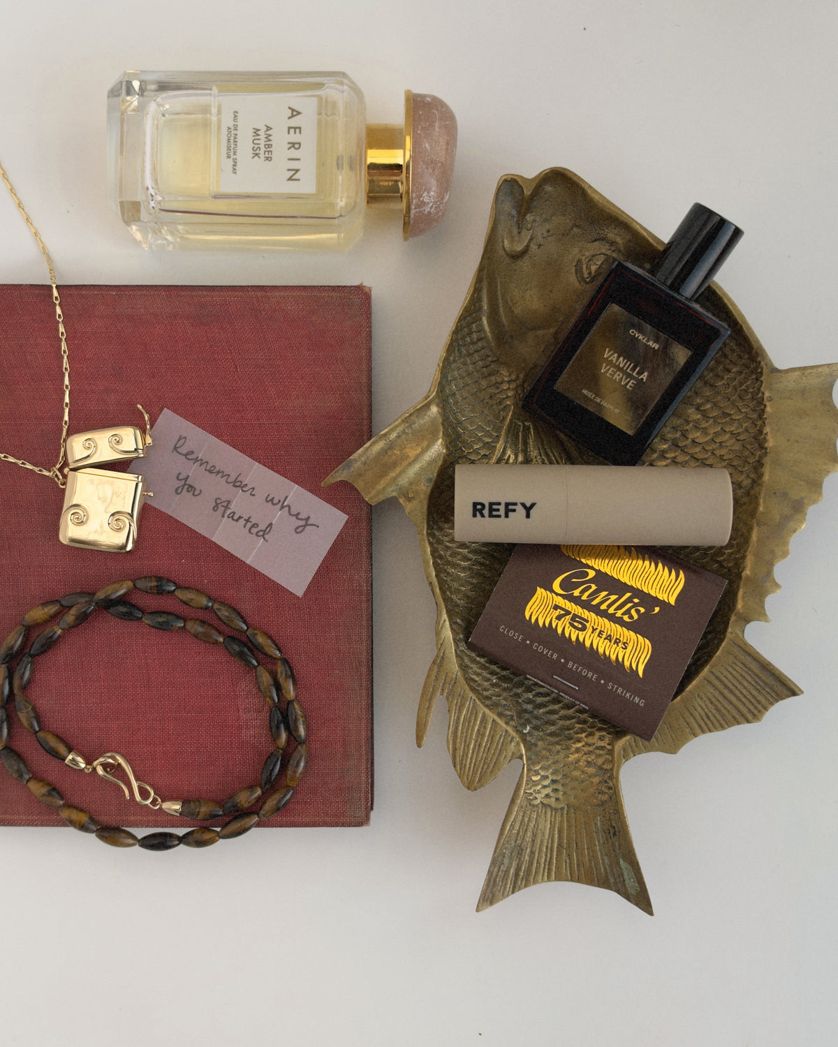 A gold-plated Sybil Vesta Case Charm on a thin chain, a beaded necklace, a red book with a note reading Remember why you started, a perfume bottle, and various beauty items arranged on a brass fish-shaped dish.