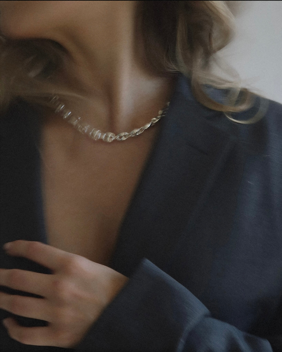 A woman wearing a dark blazer and a silver chain necklace, with her hand resting on her chest. The image is softly focused and her face is partially out of frame.