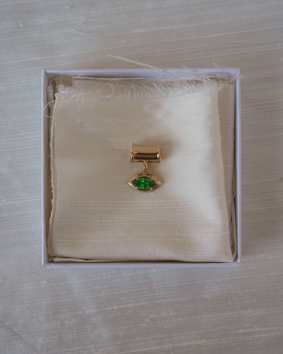 Mod + Jo Tash Charm Enhancer gold pendant necklace with a marquise-cut green stone resting on a fabric square inside a white box.