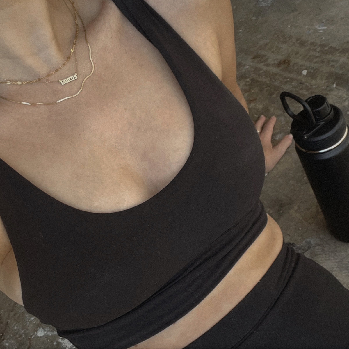 A person wearing a black sports bra and matching leggings is sitting on the ground next to a black water bottle. A gold necklace is visible around their neck. The photo is cropped to show only the torso and part of one arm.