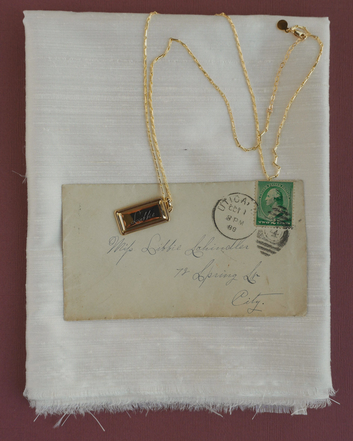 A gold rectangular Hattie Engravable Charm pendant engraved with the name Libbie resting on a vintage envelope.