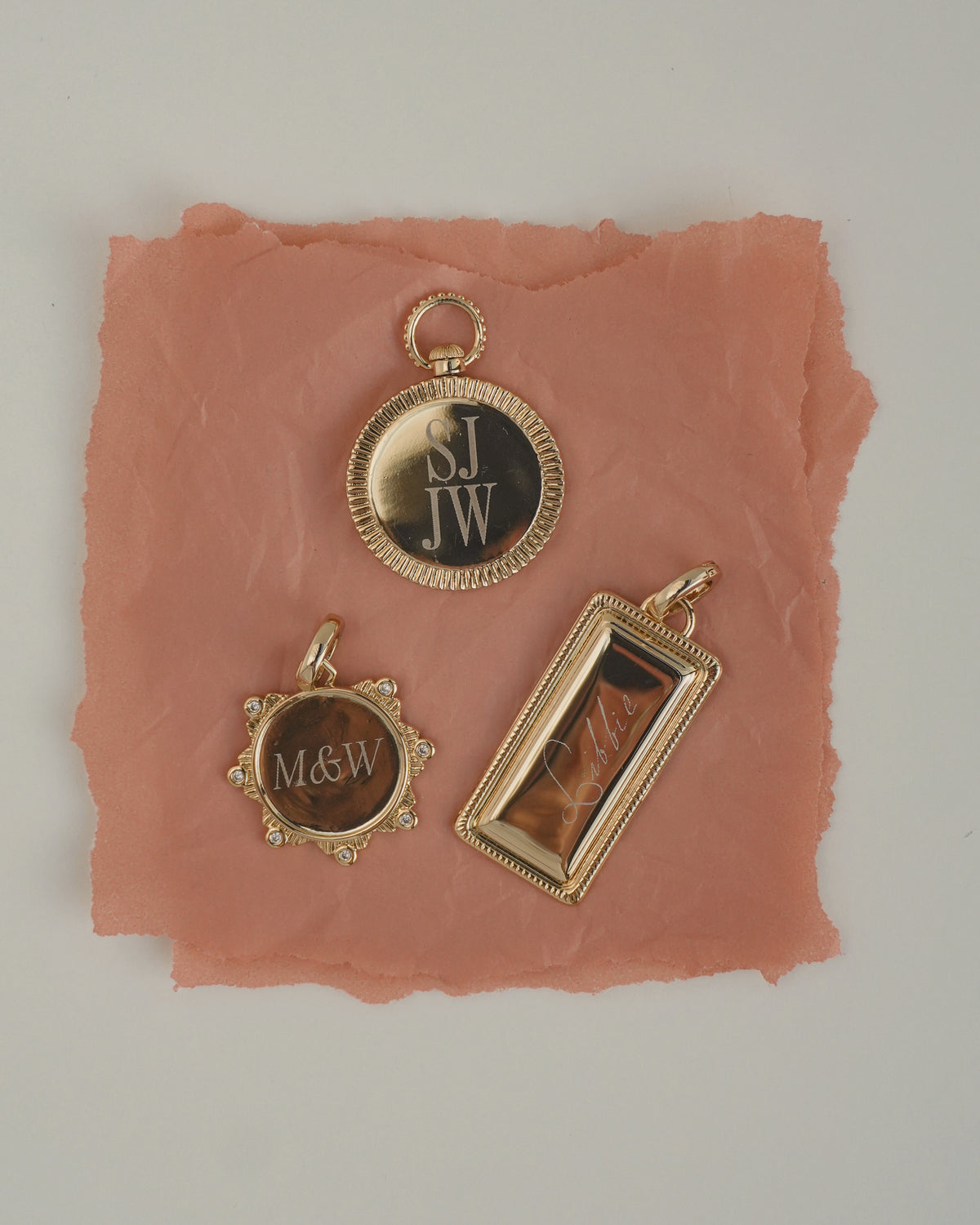 Three Remi Engravable Charm pieces on a pink paper background: a round charm marked S.J. JW, a circular cubic zirconia charm engraved with M&W, and a rectangular charm engraved with Libbie.