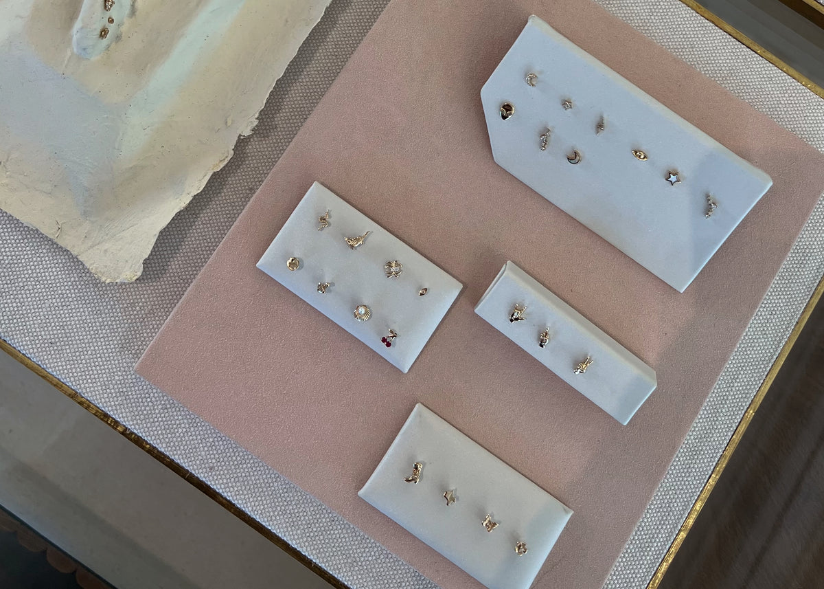 A display of various small gold and silver stud earrings arranged on white trays over a pink textured surface, with a cream-colored ceramic dish partially visible on the side.