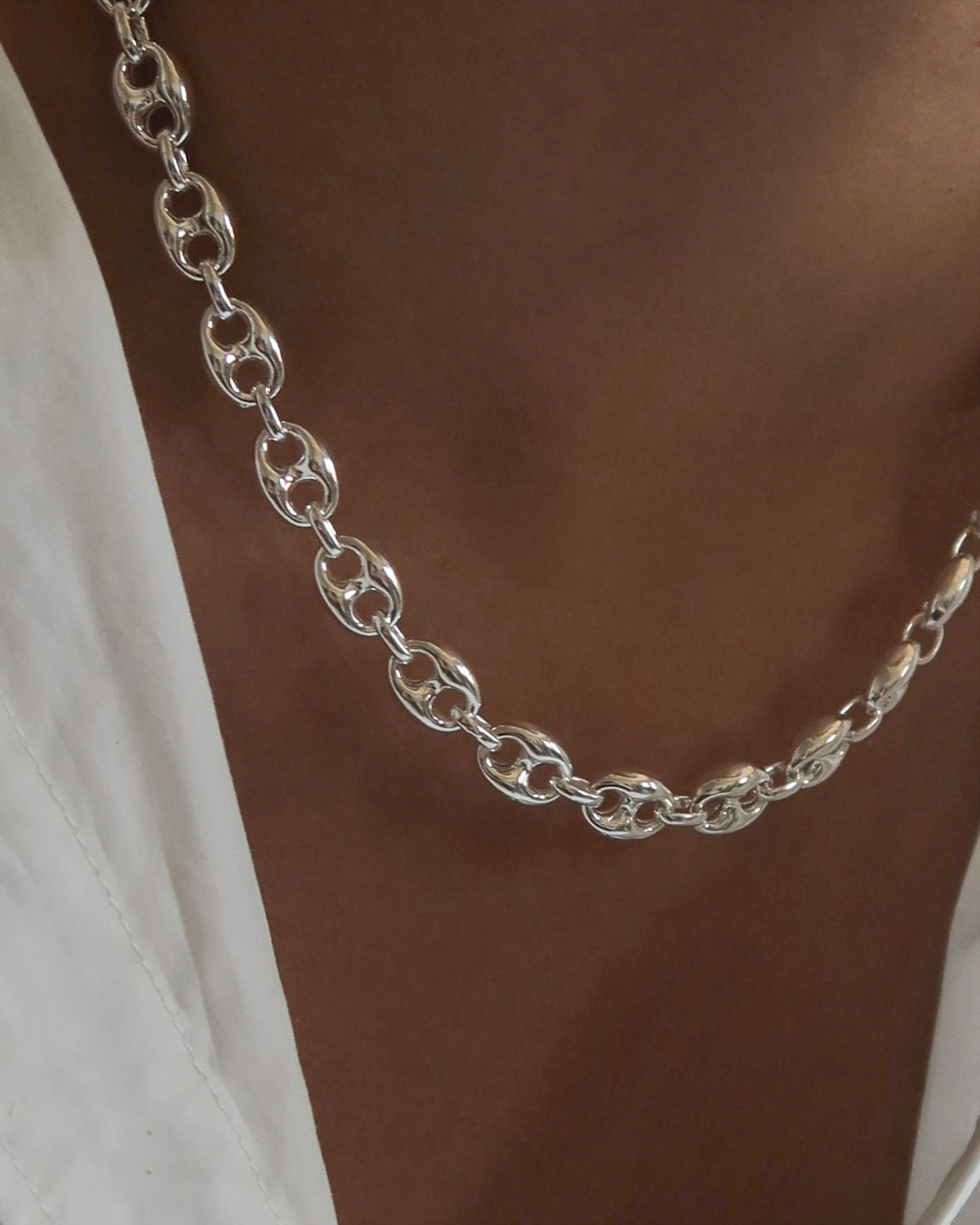 A close-up of a person wearing a chunky silver chain necklace and a white garment, shown against their brown skin.