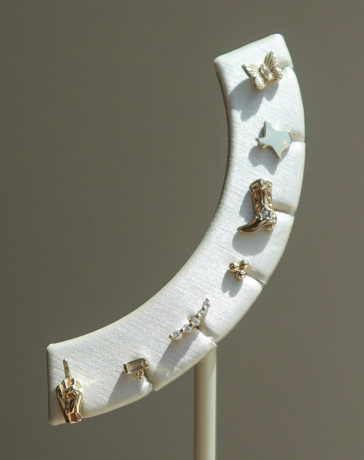 A curved white jewelry display stand holds seven single stud earrings, each with a unique design including a butterfly, star, boot, hand, and other small shapes, all in metallic finishes.