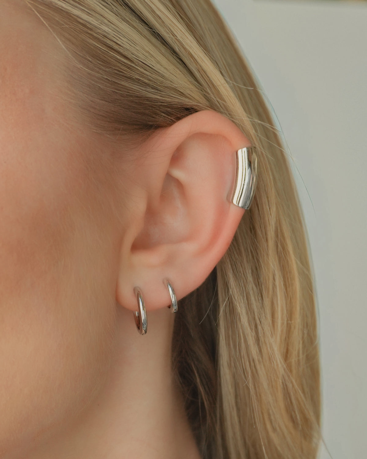A person wearing the Reneé + Lennon Set, consisting of a wide, silver helix ear cuff and two smooth huggies in the earlobe.