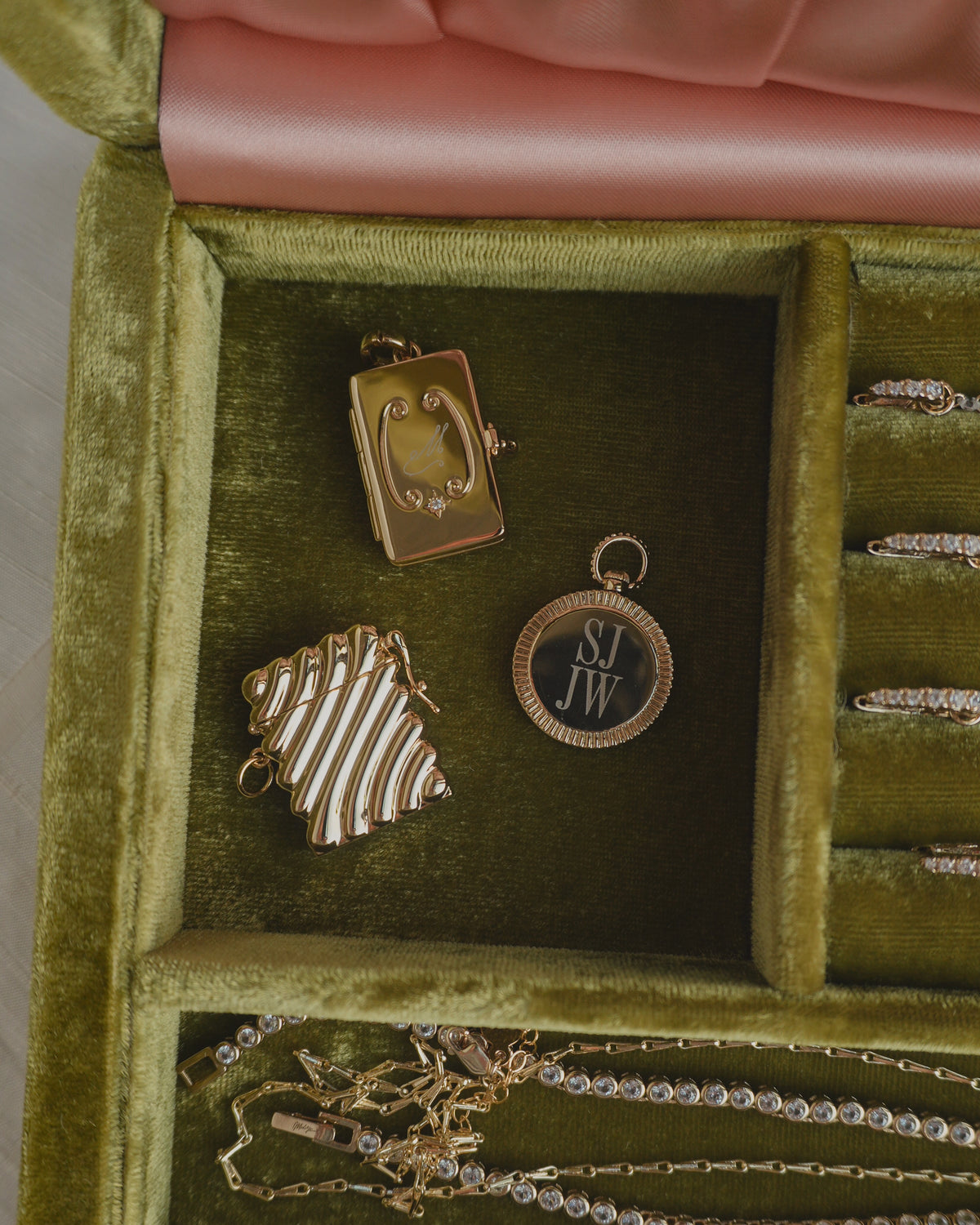 Three gold-plated jewelry charms in a green velvet box, including a Delphine Locket Charm, a striped square charm with cubic zirconia, and a circular medallion engraved with the letters SJ JW.
