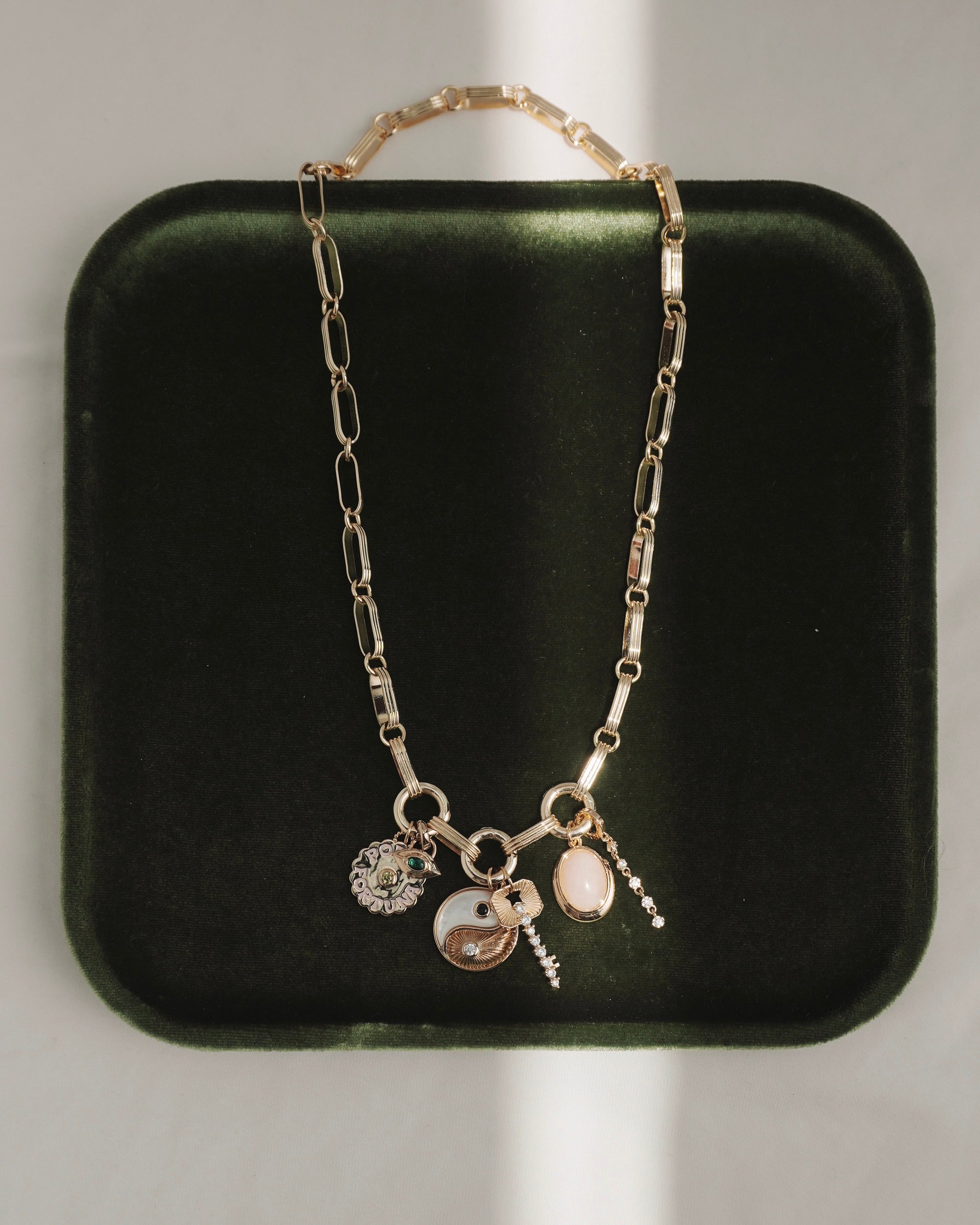 The Jagger Charm Clip Necklace by Mod + Jo, crafted in 14k gold and featuring four unique charms—a yin-yang, a flower, an oval stone, and a petite hanging pendant—is elegantly displayed on a dark green velvet tray with sunlight creating gentle shadows.