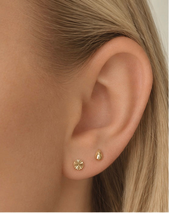 A pair of gold Dela + Uma Set stud earrings in an earlobe, featuring a fluted sunburst design and a smooth, teardrop stud.