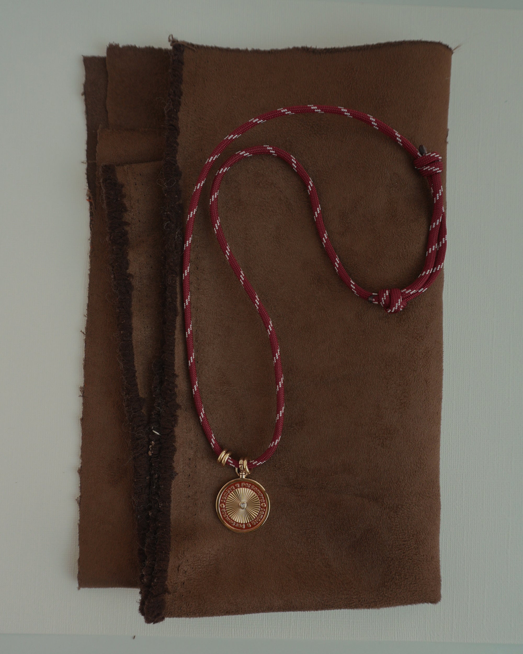 The Tommy Cord Necklace by Mod + Jo, featuring a vibrant red paracord and a round gold pendant with subtle gold plate accents, is displayed atop a folded piece of brown suede fabric.