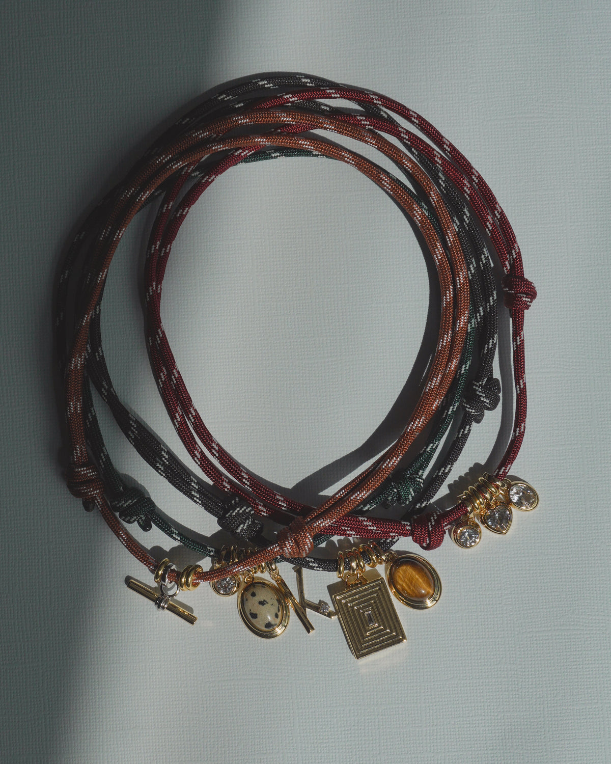 Three multicolored Tommy Cord Necklaces by Mod + Jo, adorned with gold and gemstone charms, are arranged in overlapping circles on a light textured surface with sunlight casting delicate shadows. Gold-plated spacers add an elegant touch to each necklace, enhancing the vibrant look of these lively accessories.