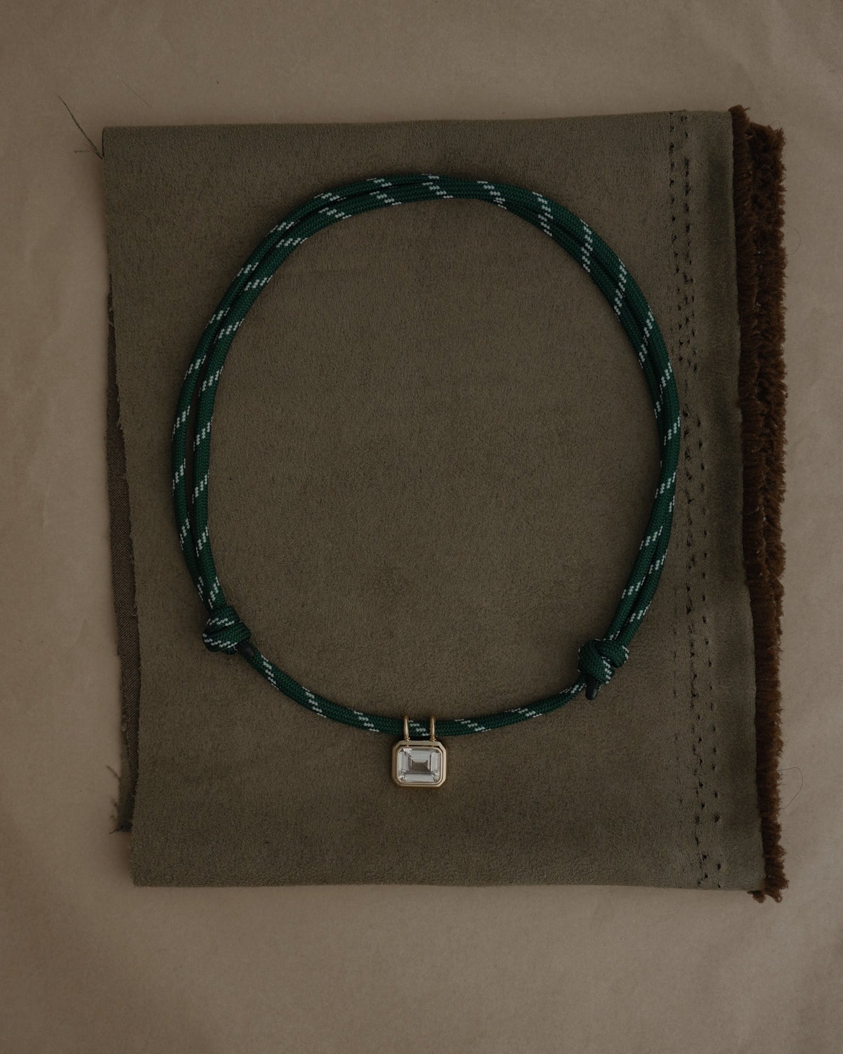 The Mod + Jo Tommy Sapphire Pendant Necklace features an adjustable green and white paracord with a small square gold bezel-set sapphire, showcased on a folded piece of brown fabric.