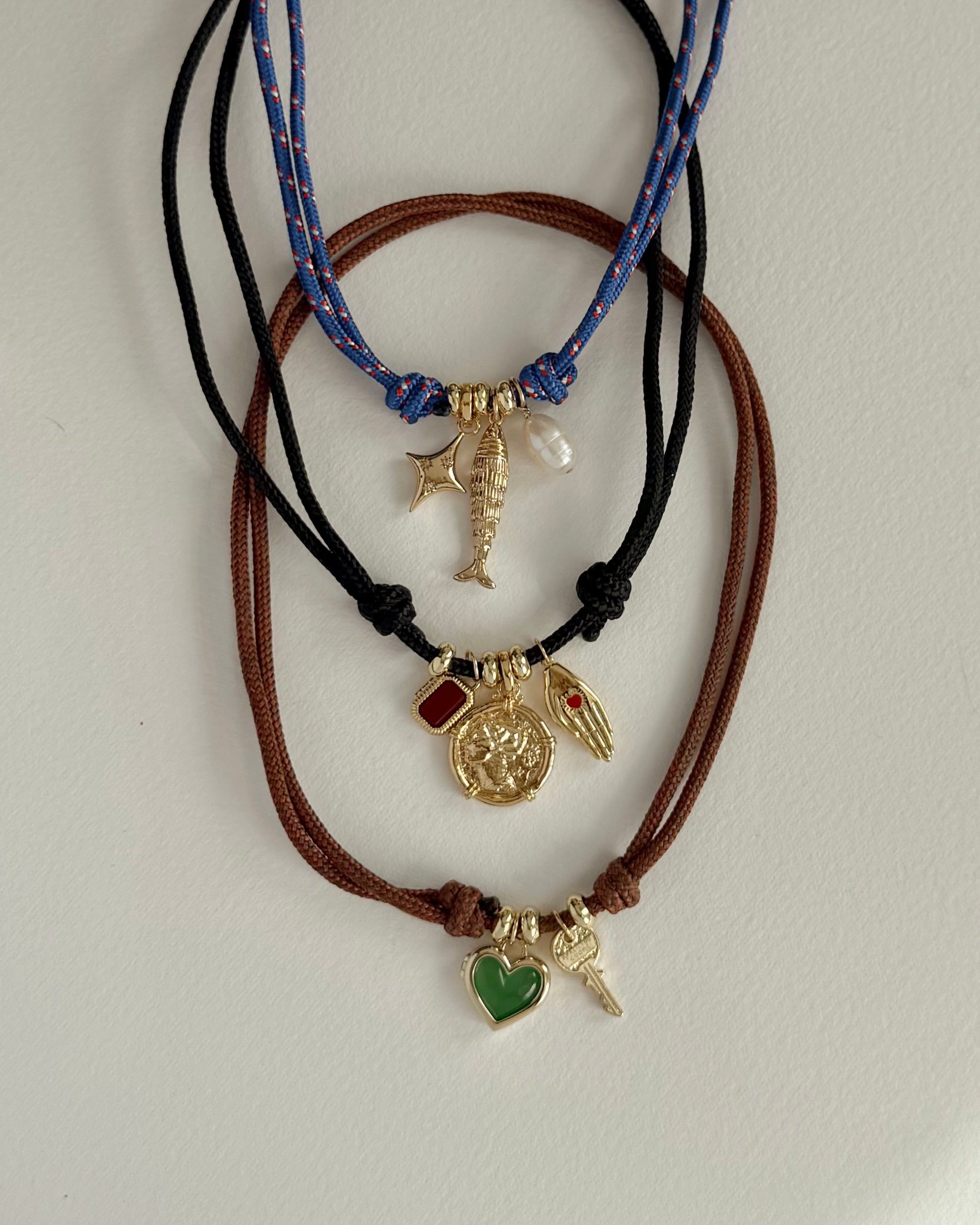 Three Mod + Jo cord bracelets on a white surface: the top with gold fish, pearl, and bar charms; the middle with genuine gemstone charms—a red stone, sun, and wing; the bottom features a Davie Heart Birthstone Locket Charm.