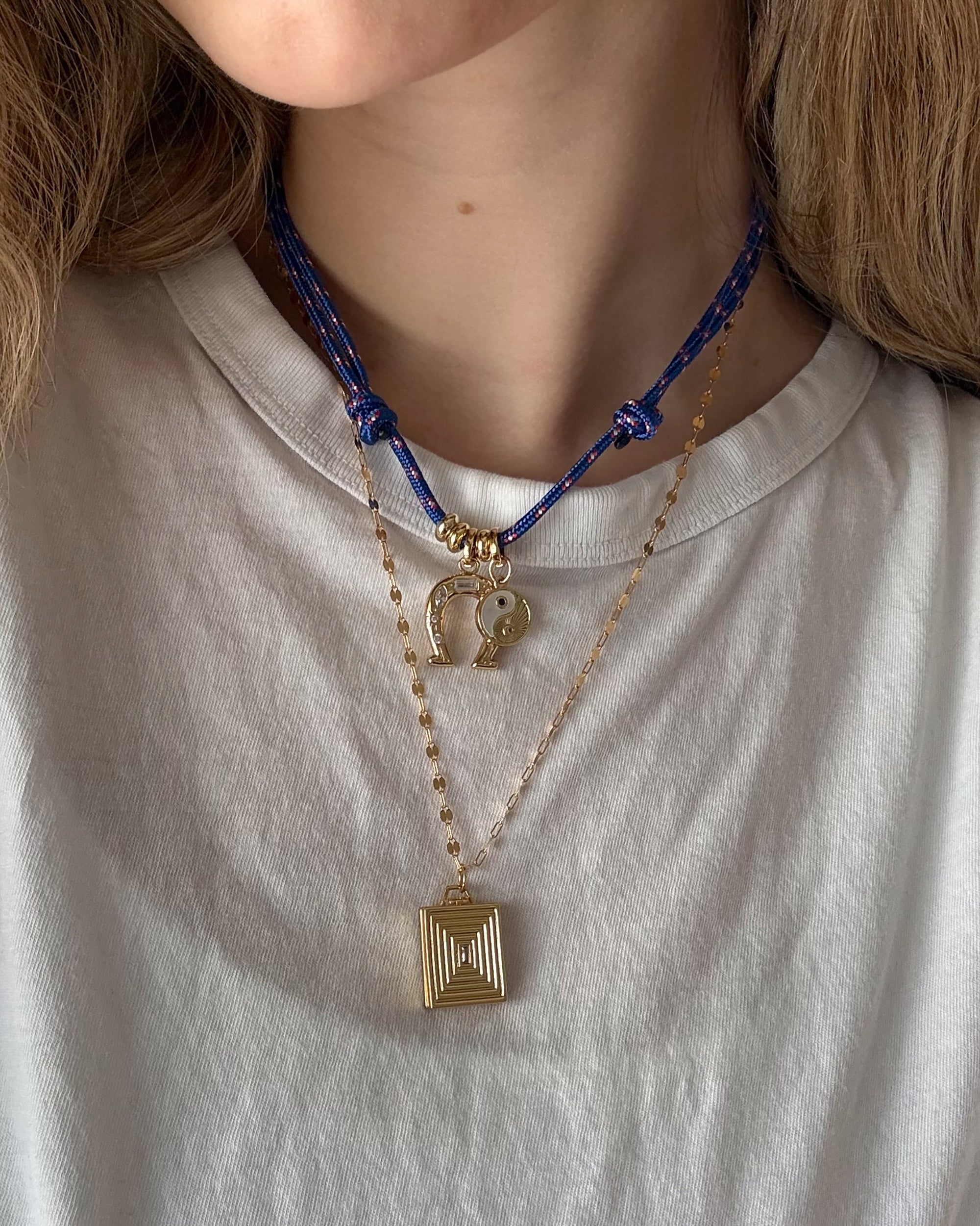 A person in a white t-shirt wears two necklaces: a bold blue cord necklace with gold charms, including a yin-yang, and Mod + Jo’s Inez Locket Charm—a delicate 14k gold plated chain with a square, textured pendant.