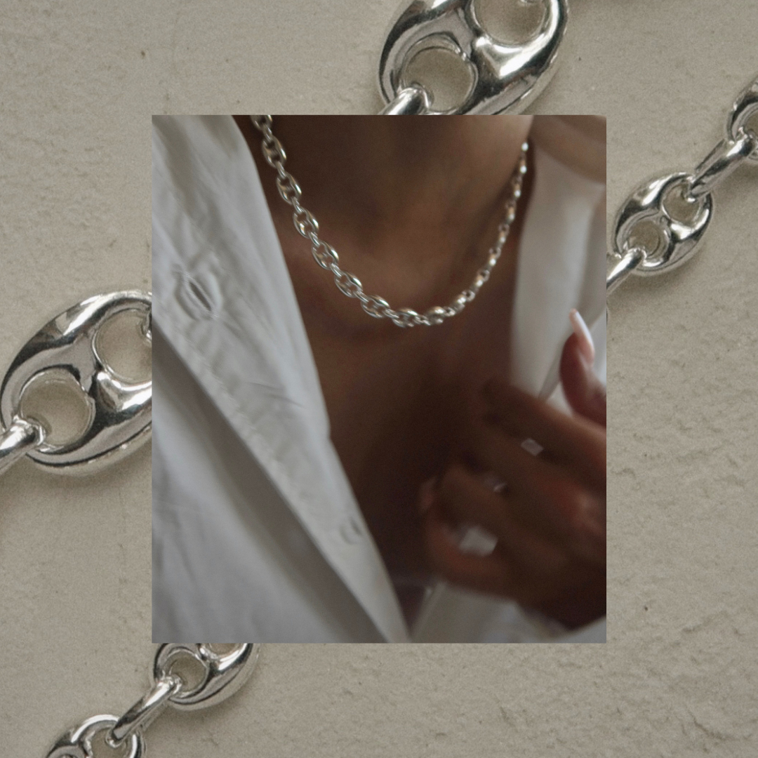 A close-up of a person wearing a thick silver chain necklace over an open white shirt. The background features a pattern of large silver chain links on a beige surface. The persons hand is near their collar.
