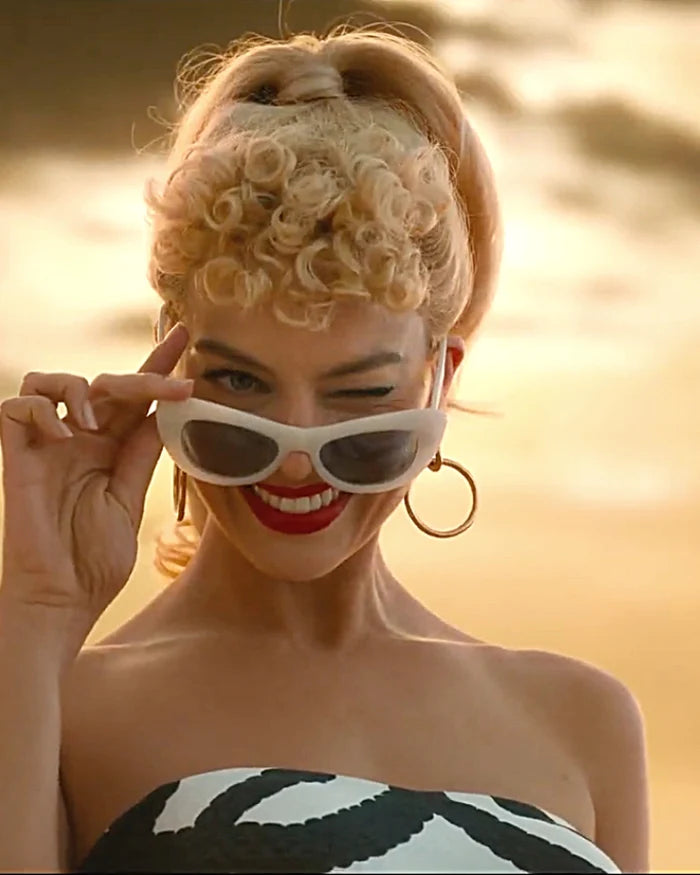 A woman with blonde curly hair in a ponytail winks and smiles, holding white cat-eye sunglasses. She wears red lipstick, gold hoop earrings, and a strapless top, with a golden sky in the background.