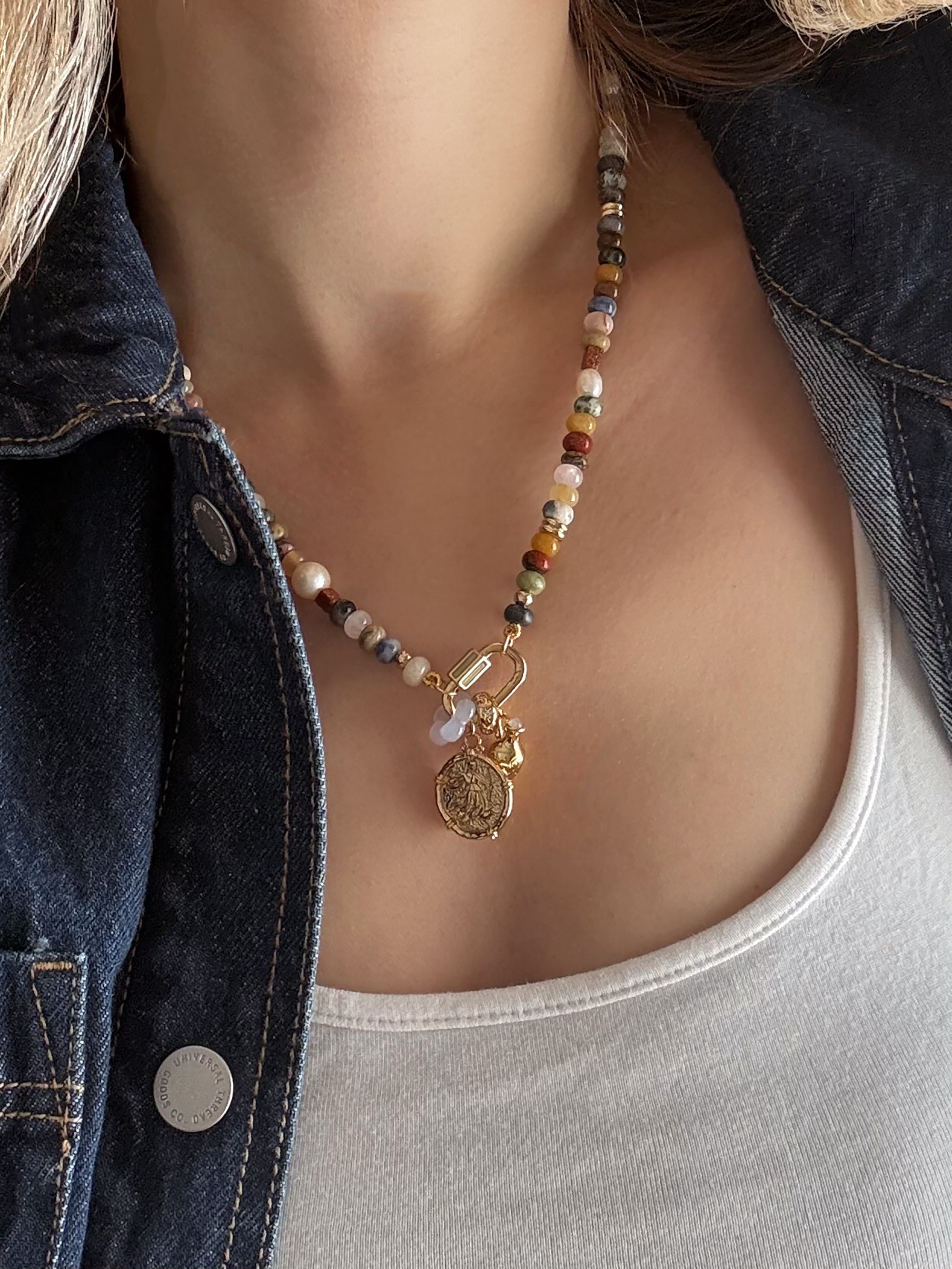 A person styled in a white top and dark denim jacket wears the Mod + Jo Andy Gemstone Enhancer Chain, a 14k plated necklace featuring colorful beads, gold coin charm, and a small gemstone.