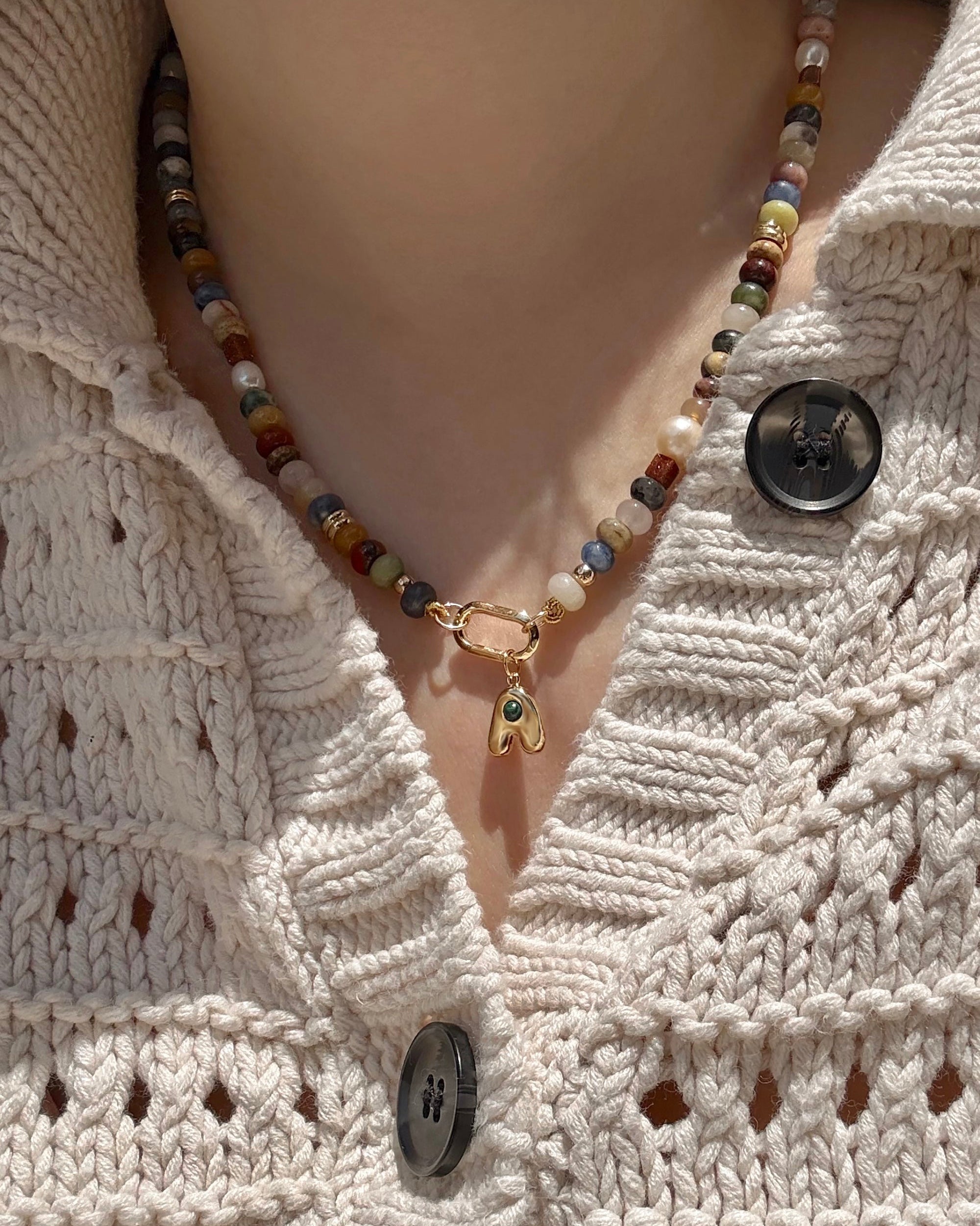 A close-up shows a person in a beige knitted sweater with large black buttons, wearing the Mod + Jo Andy Gemstone Enhancer Chain—a beaded necklace with 14k plating and a small gold eye pendant.