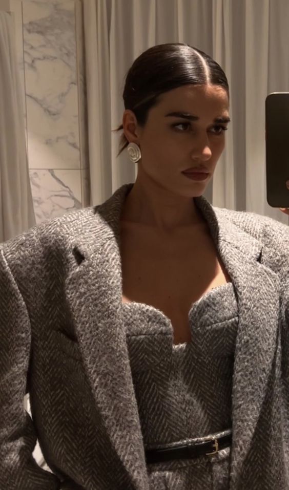 A woman with slicked-back dark hair wears a grey herringbone blazer and matching strapless top, accessorized with large round earrings. She takes a mirror selfie in a room with white curtains and marble walls.