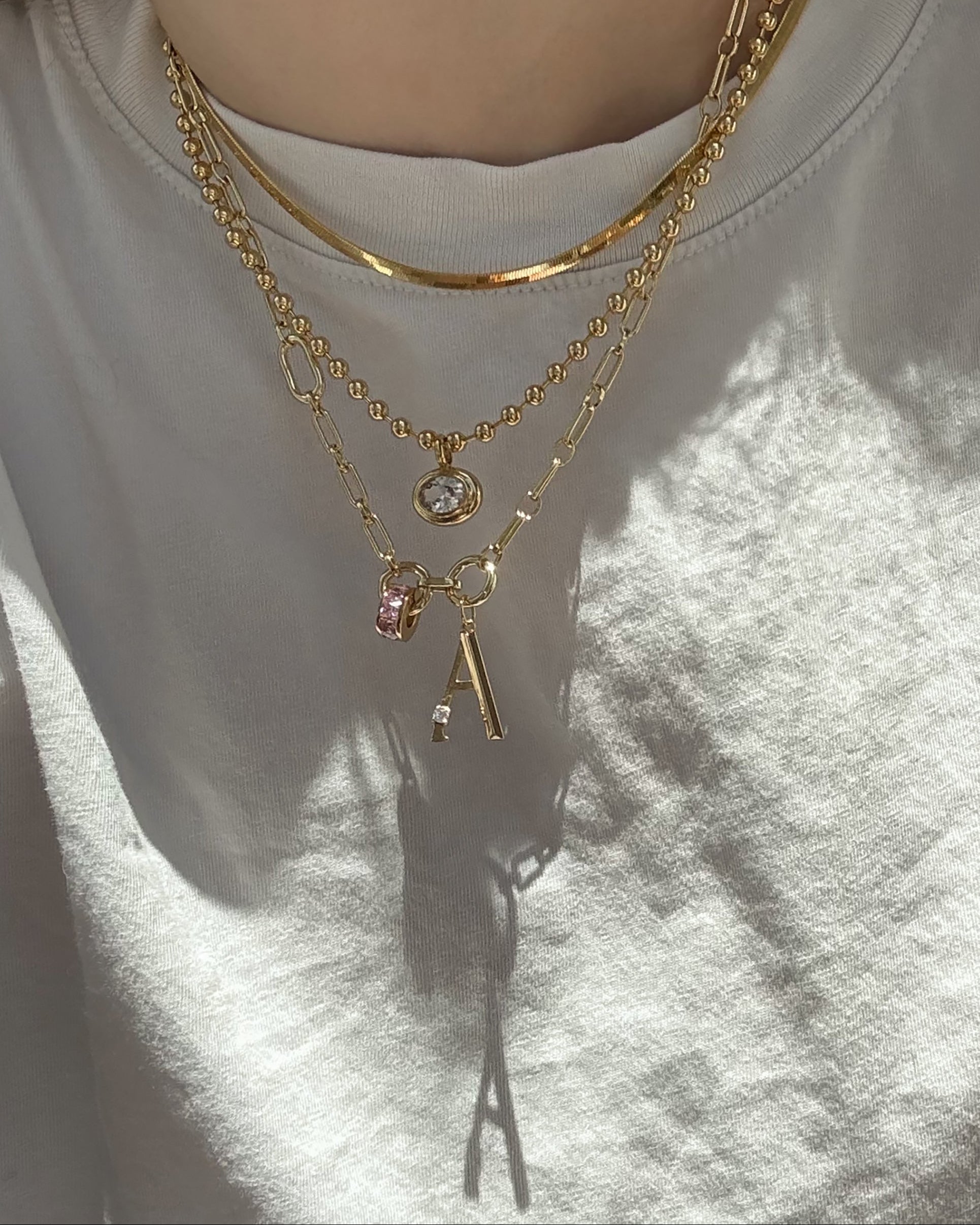 A person wears a white shirt and three layered gold plated necklaces, including a chain, a pendant with a clear stone, and the Mod + Jo Eloise Letter Charm featuring an initial A and pink bead. Sunlight casts shadows on the shirt.