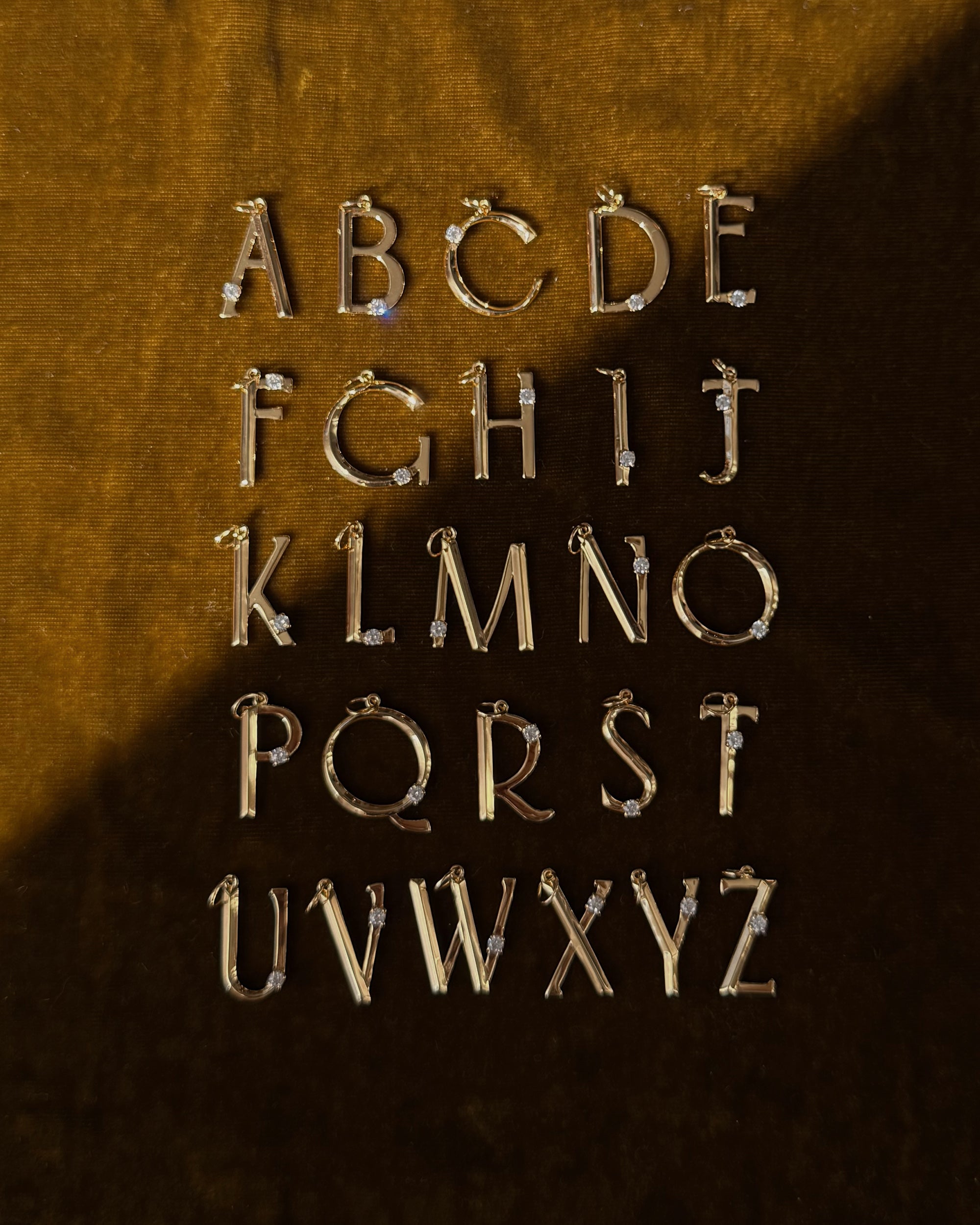 The Eloise Letter Charm by Mod + Jo features gold alphabet pendants with small gemstones, elegantly arranged from A to Z on dark brown velvet—the ideal personalized charm for your necklace or initial collection.