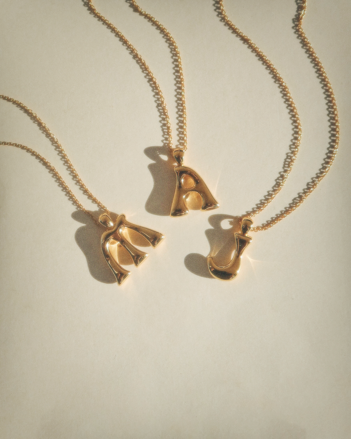 Three gold necklaces with letter pendants arranged on a cream background. The pendants form the letters M, A, and J. The chains are delicate and the lighting casts soft shadows.