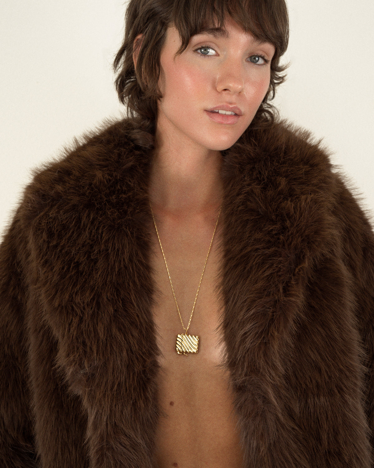 A person wearing a gold Caspia Vesta Case Charm necklace, featuring a square, textured, rippled design.