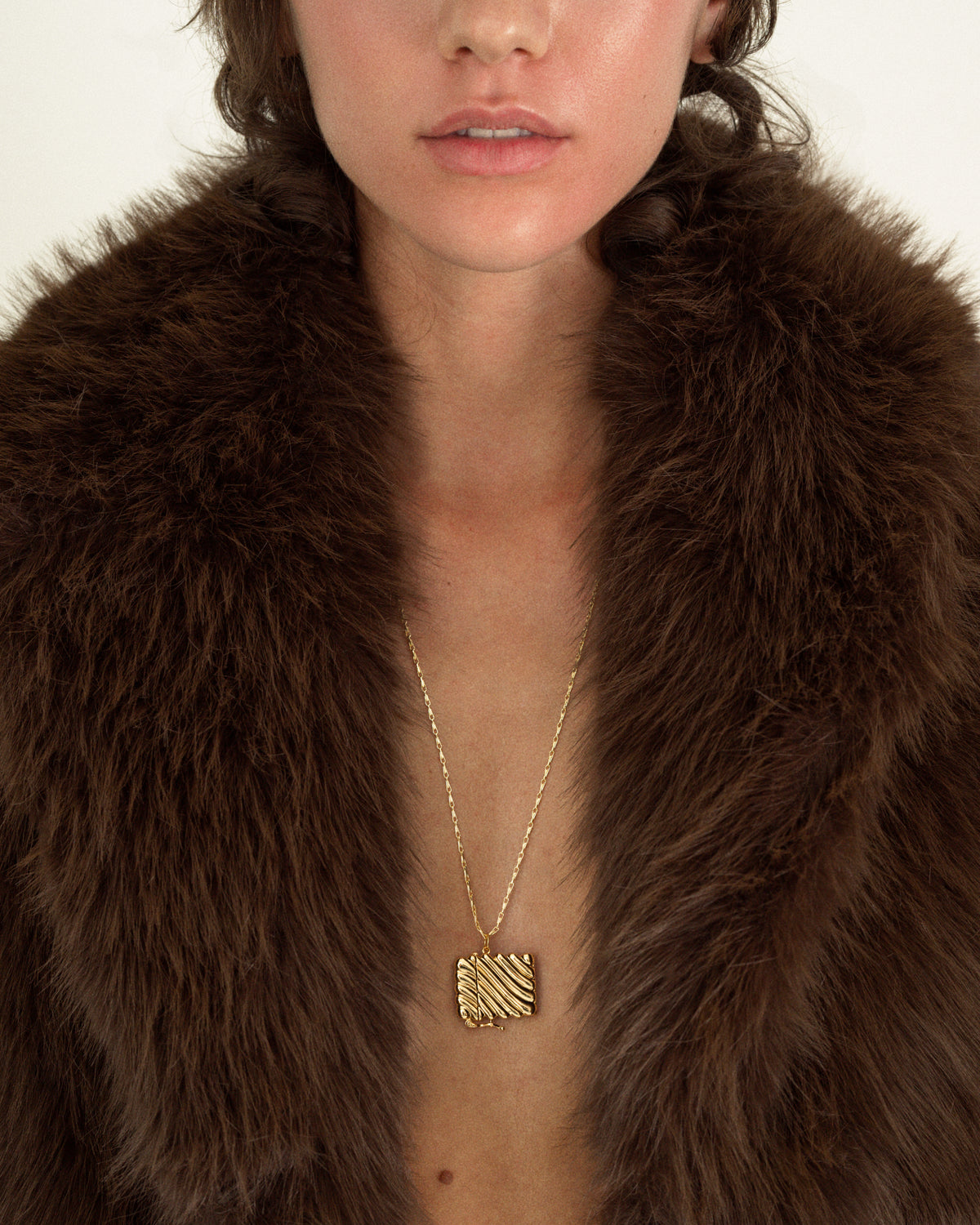 A person wears a delicate gold plate Caspia Vesta Case Charm featuring a square, textured red jasper pendant over a brown faux-fur coat.